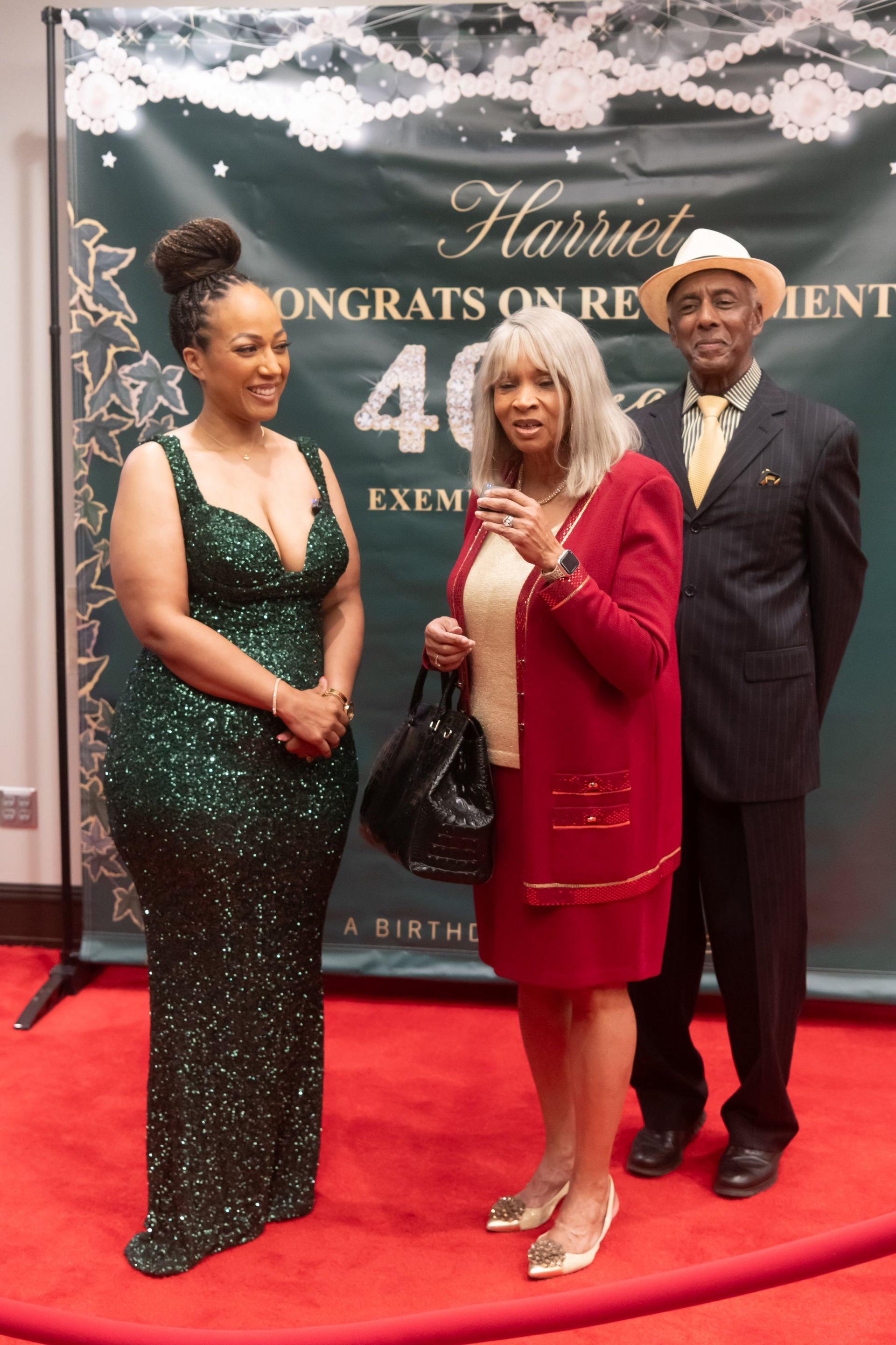 Three people posing on a red carpet at a 40th anniversary event, with a dark backdrop and floral decorations.