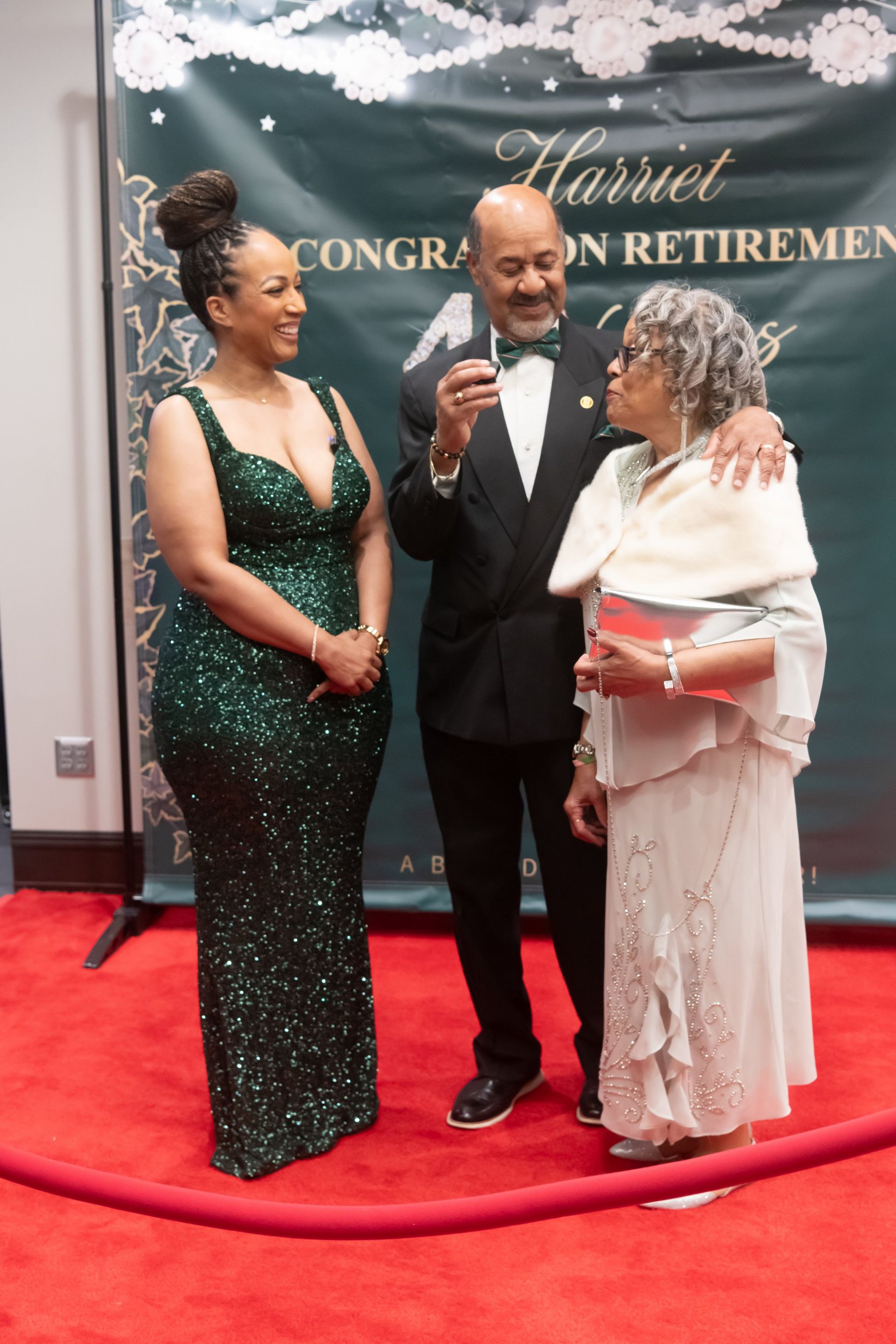 Three people in formal wear on a red carpet at a retirement celebration, holding microphones and a plaque.