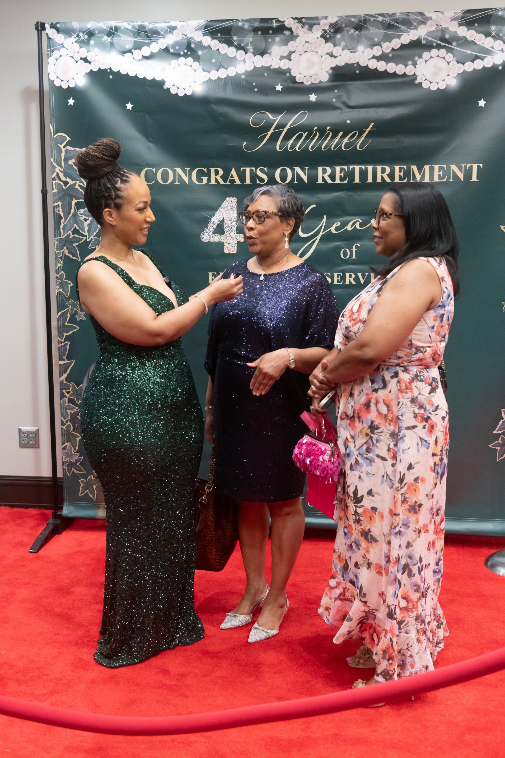 Three women in formal dresses chatting on a red carpet at a retirement celebration backdrop