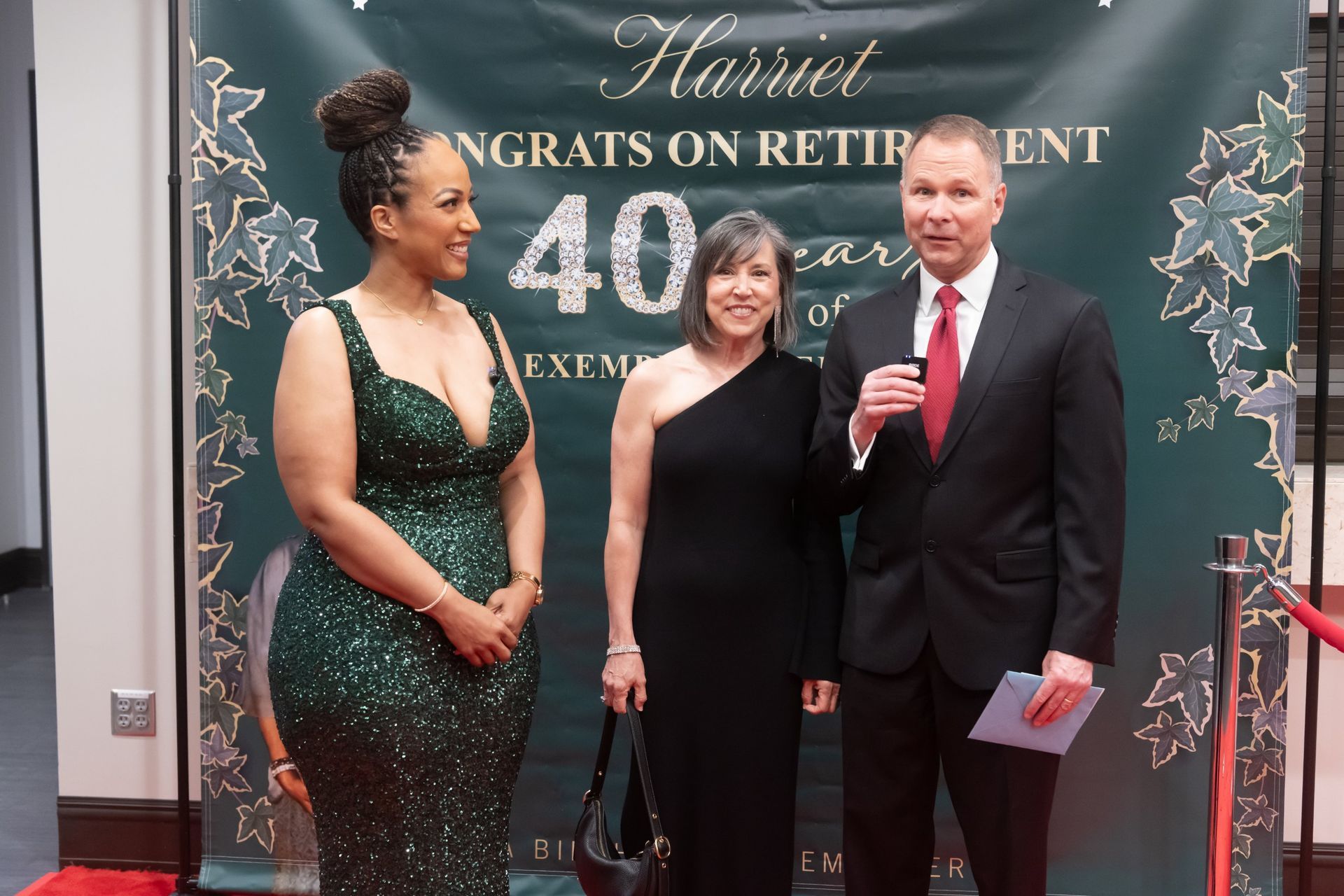 Three people posing at a retirement celebration backdrop, one holding a microphone and papers.