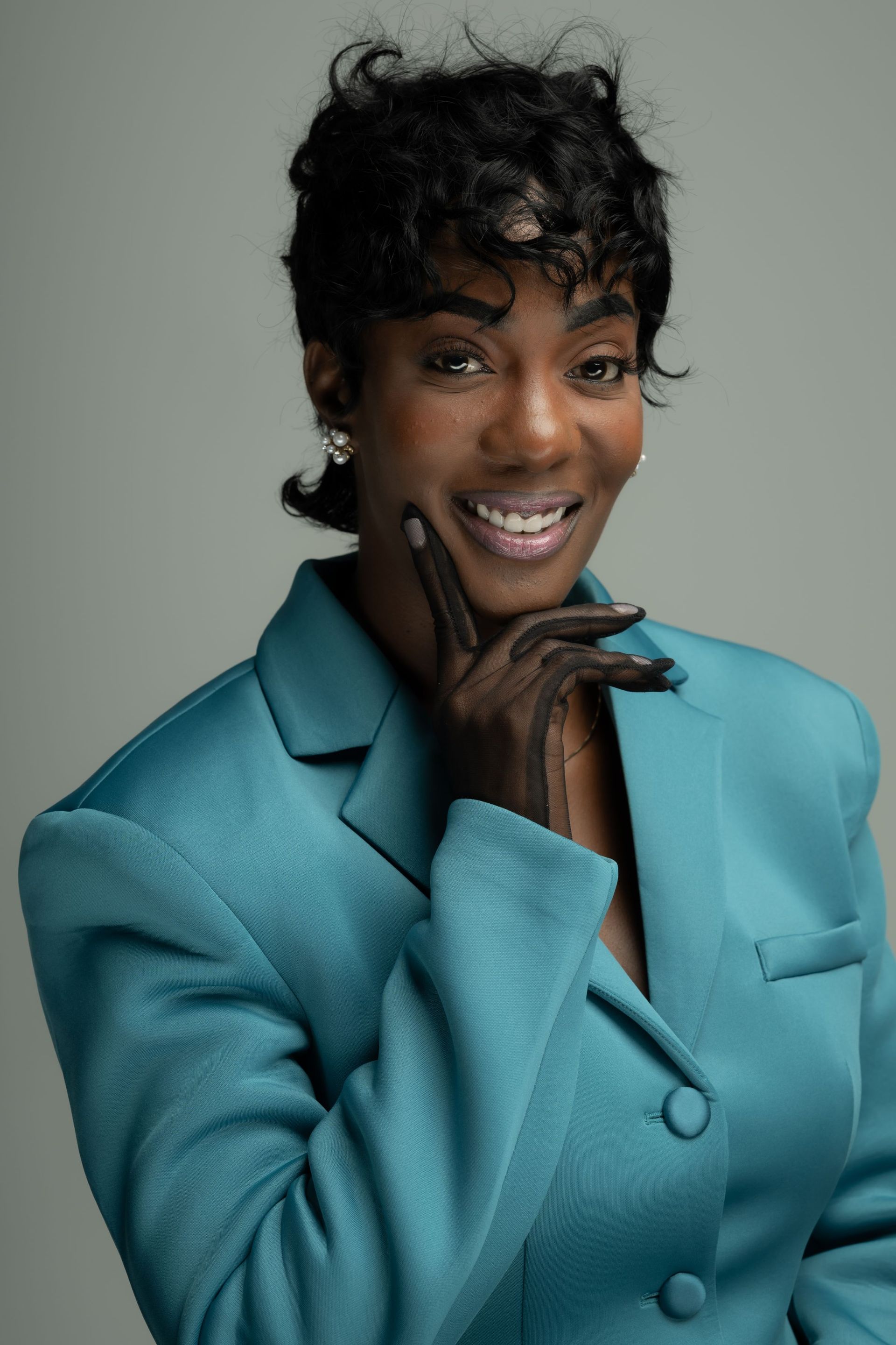 Smiling person in a teal blazer posing with hand under chin against a gray background