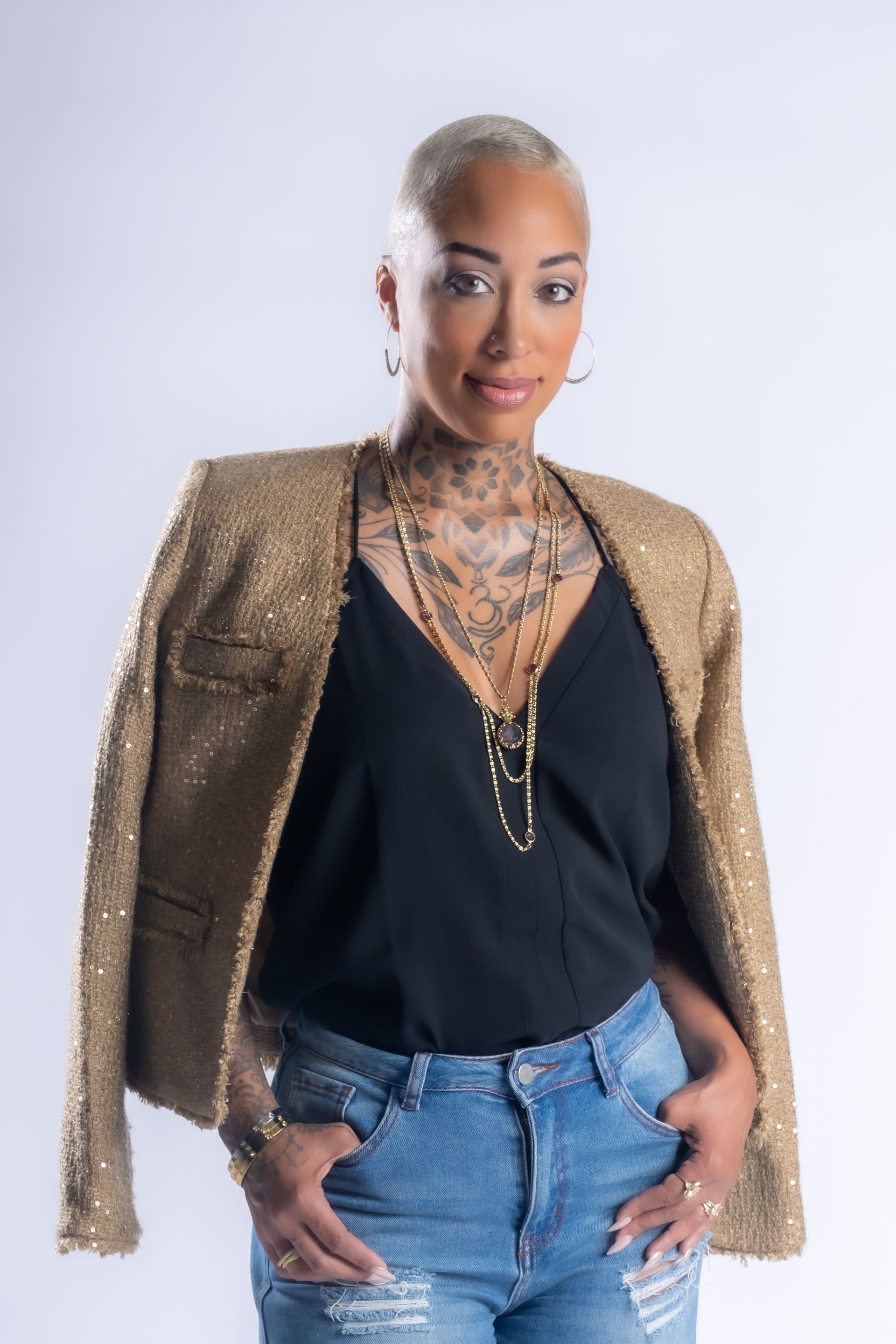 Person posing in a gold jacket, black top, and jeans against a light background.