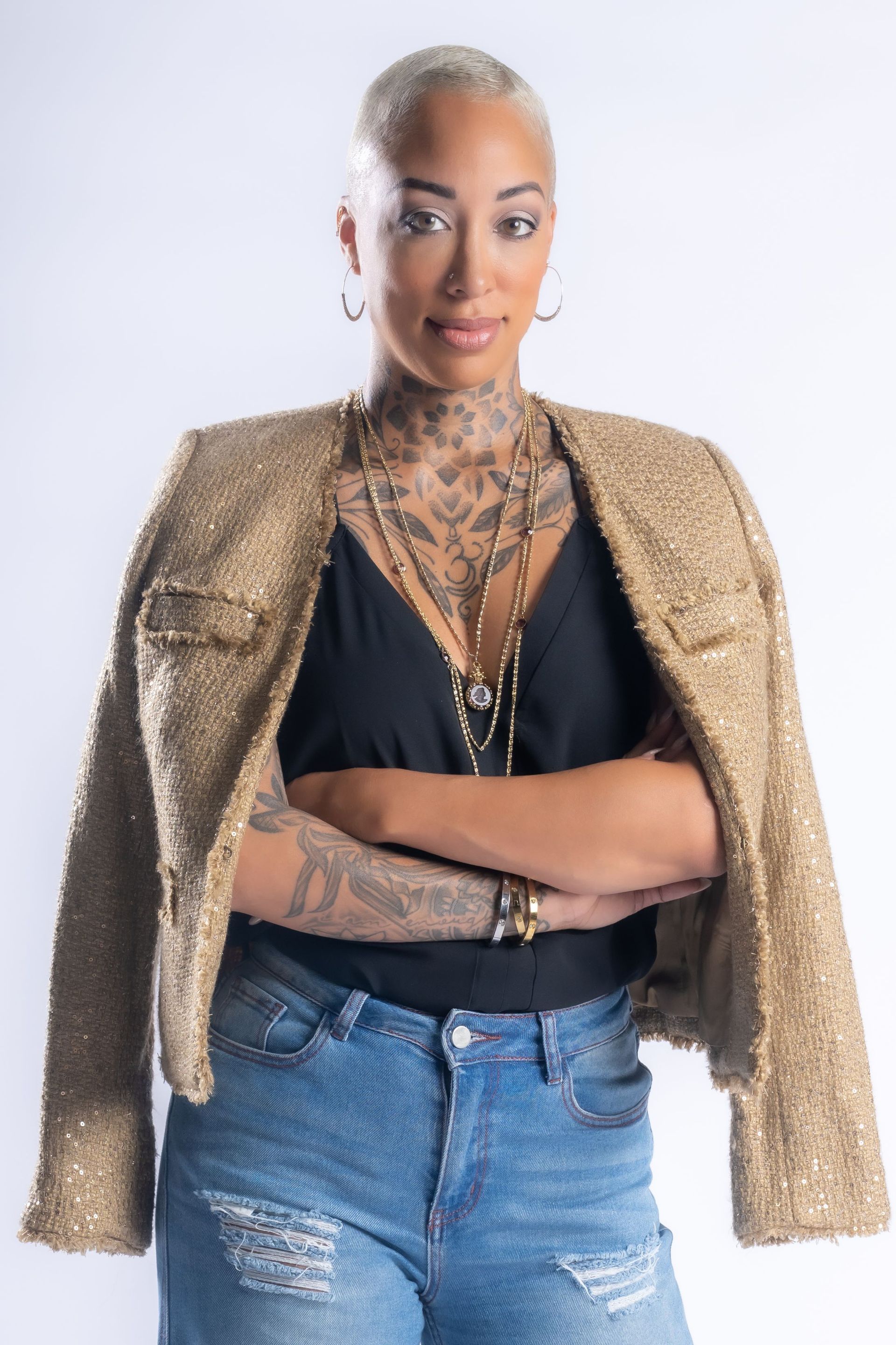 Woman posing in a gold jacket, black top, and blue jeans against a light background