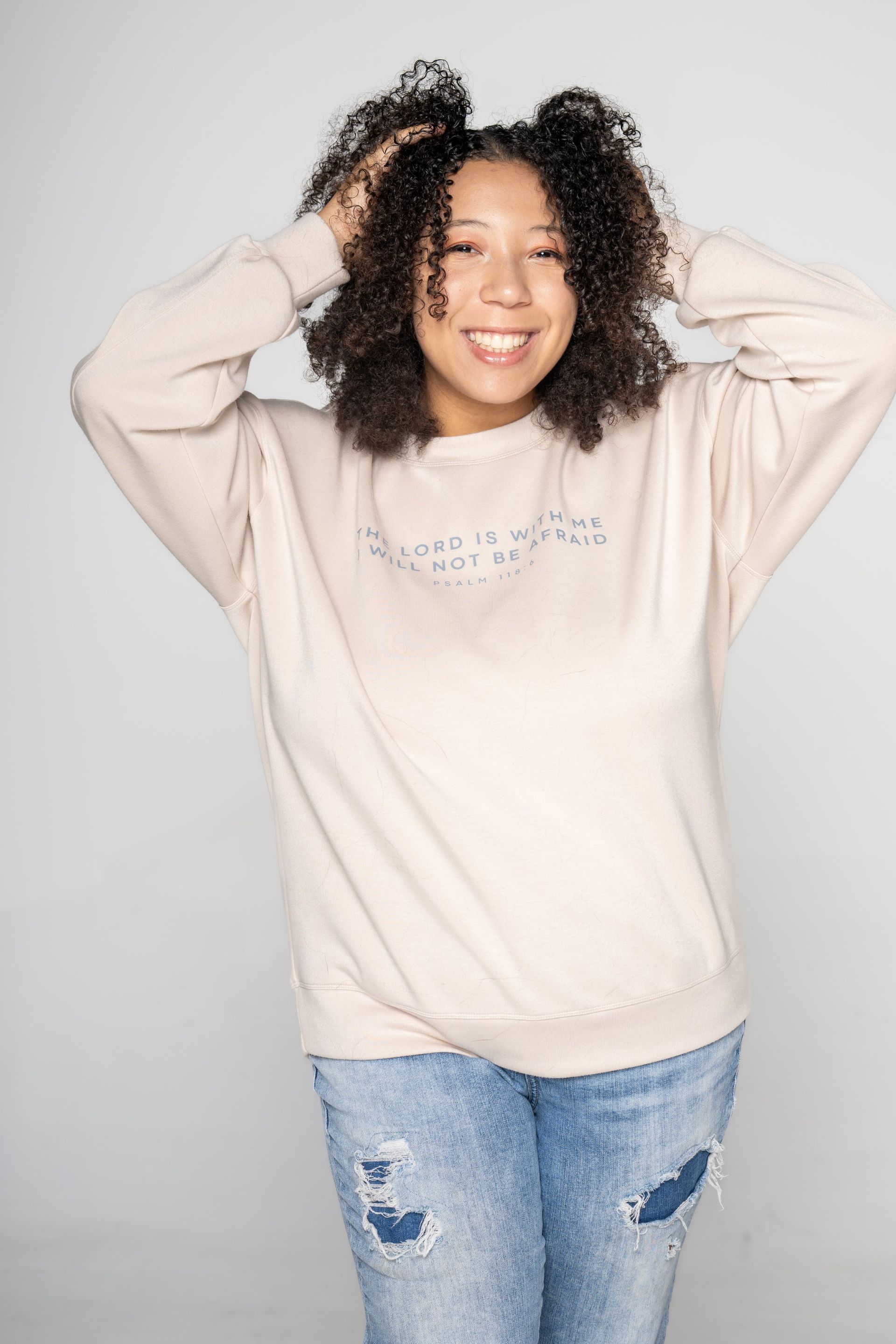 Woman smiling with hands in curly hair, wearing a beige sweater and ripped jeans against a light background