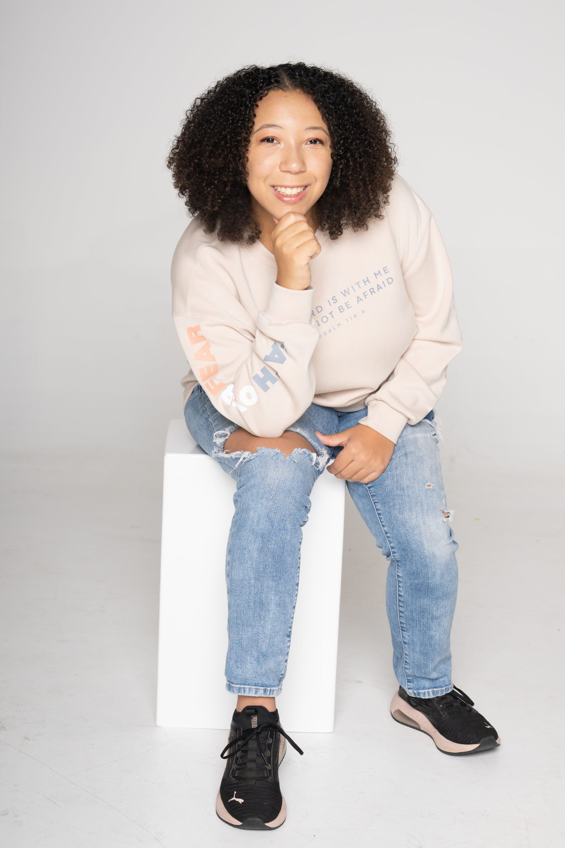 Person sitting on a white block, wearing a beige sweatshirt, ripped blue jeans, and black sneakers, smiling at camera
