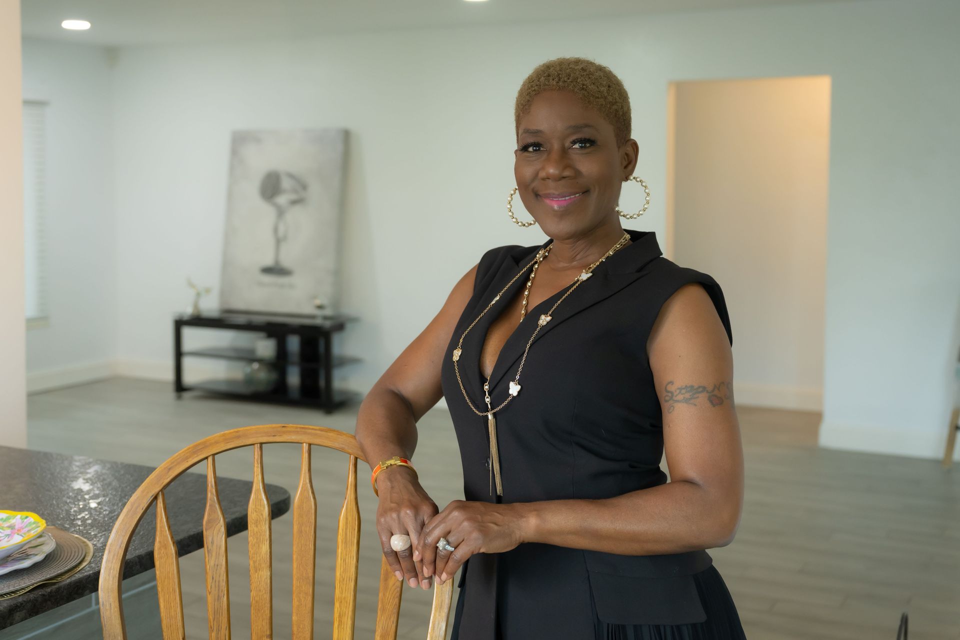 Woman standing indoors beside a wooden chair, smiling in a bright, minimalist room.