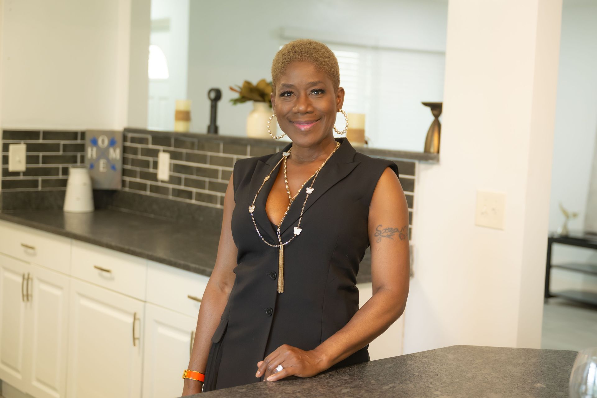 Woman standing in a bright kitchen, smiling beside a dark countertop and white cabinets.