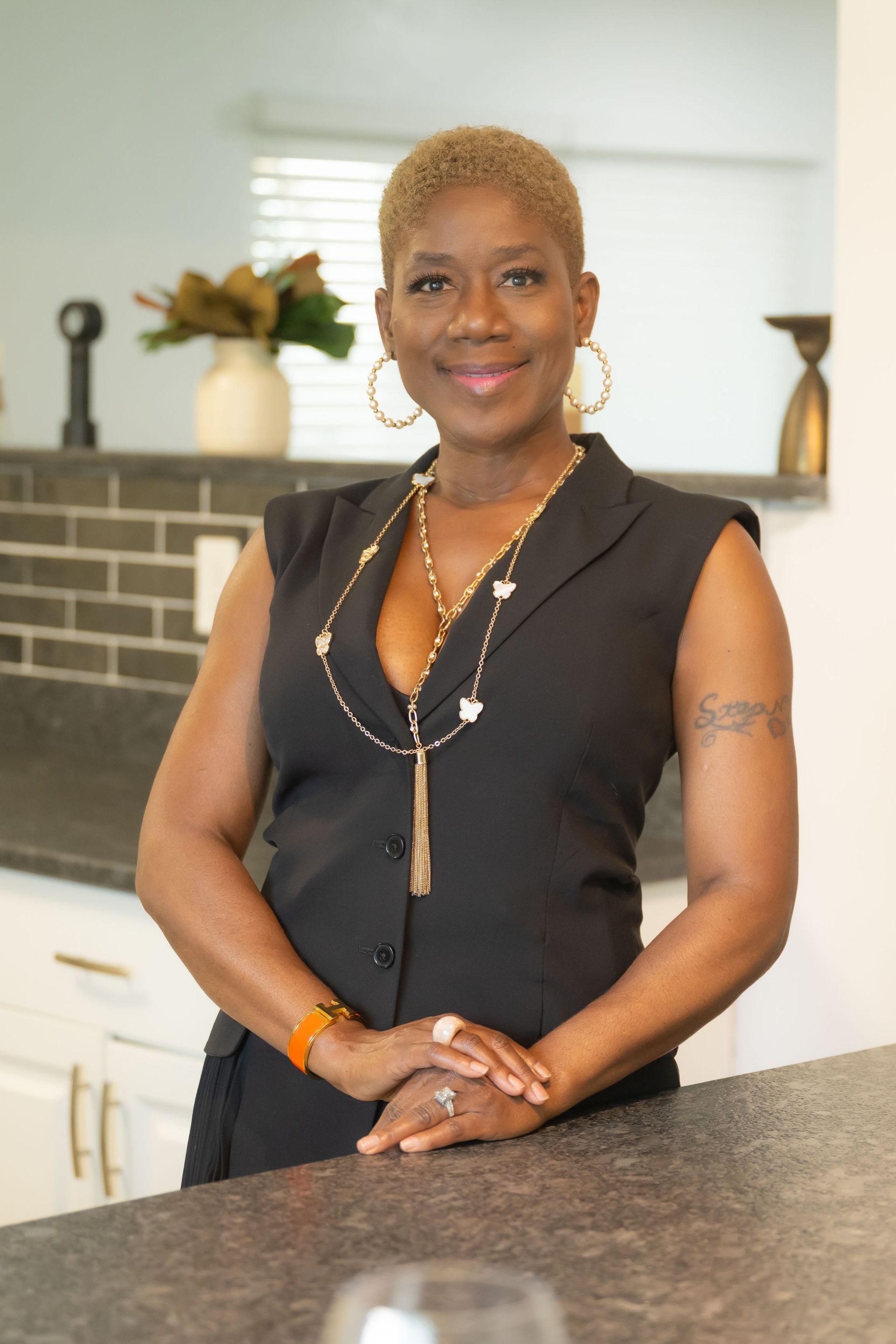 Woman standing behind a counter in a bright kitchen, smiling with hands clasped.