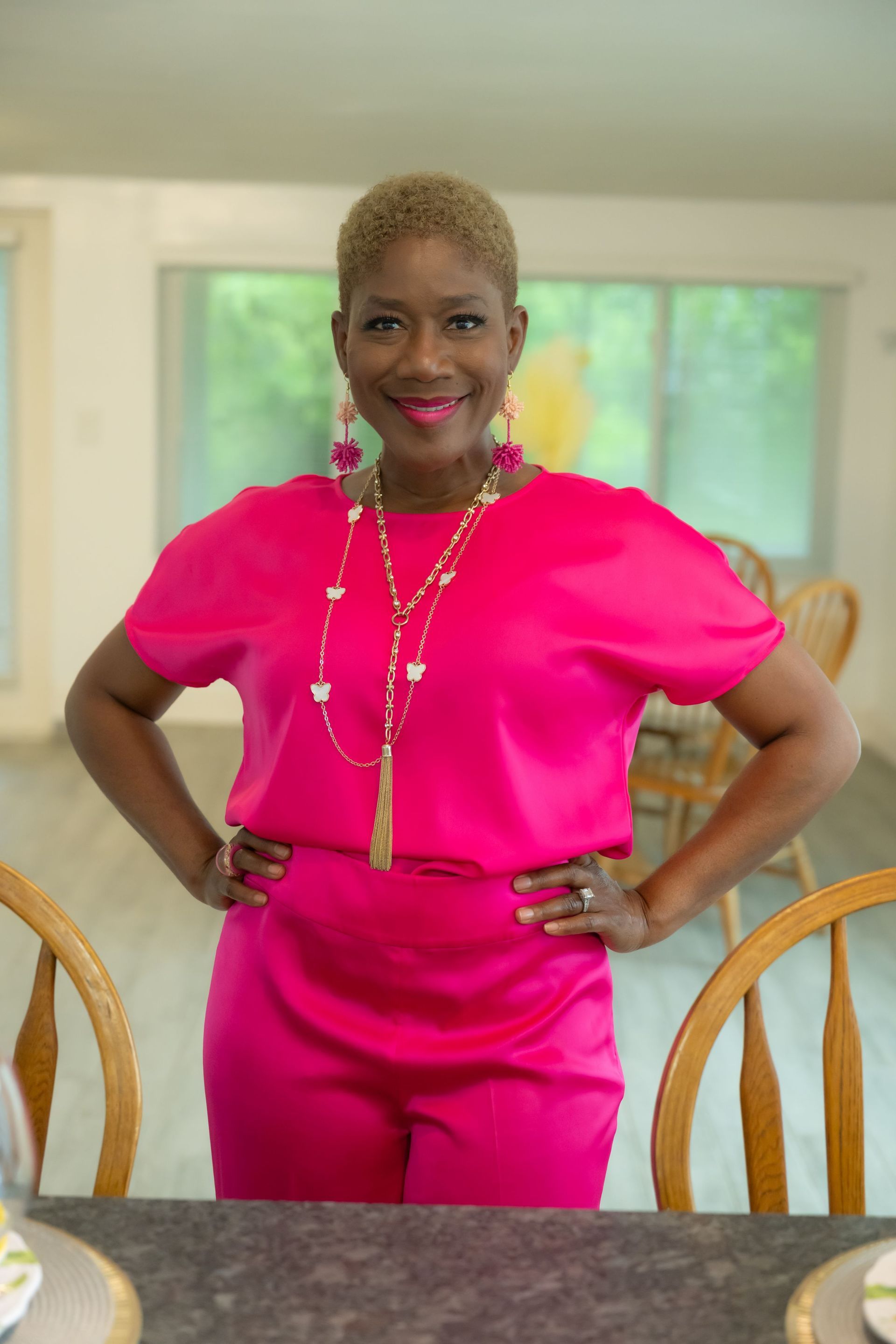 Woman in bright pink outfit standing with hands on hips in a sunlit dining room