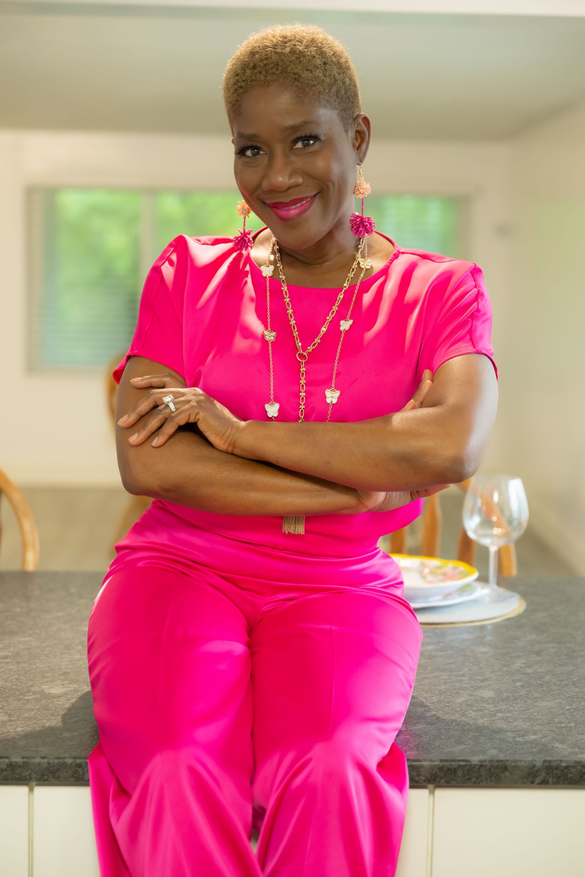 Woman in bright pink outfit leaning on a kitchen counter, smiling with arms crossed.