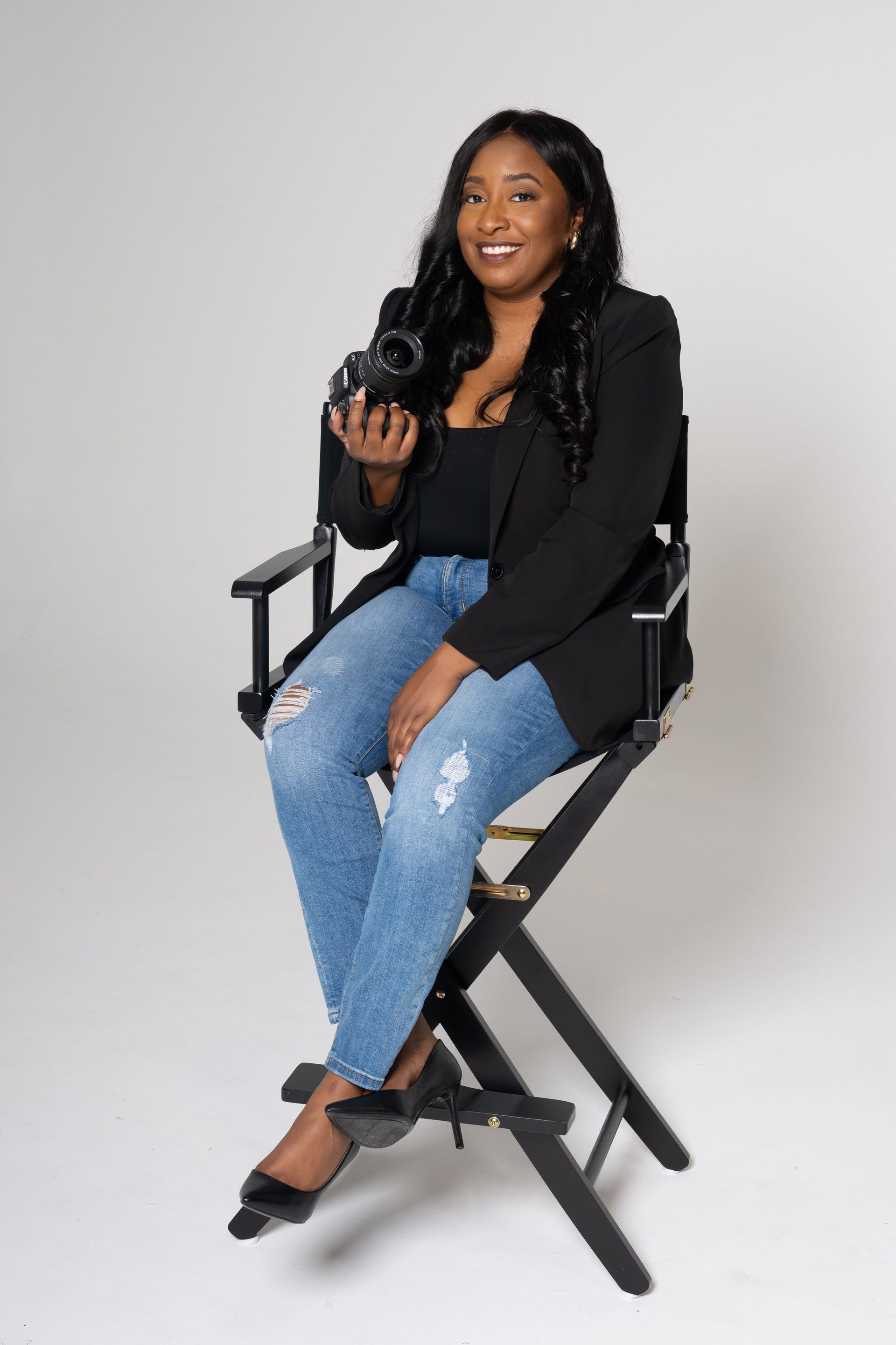 Woman seated on a director’s chair holding a camera against a white background