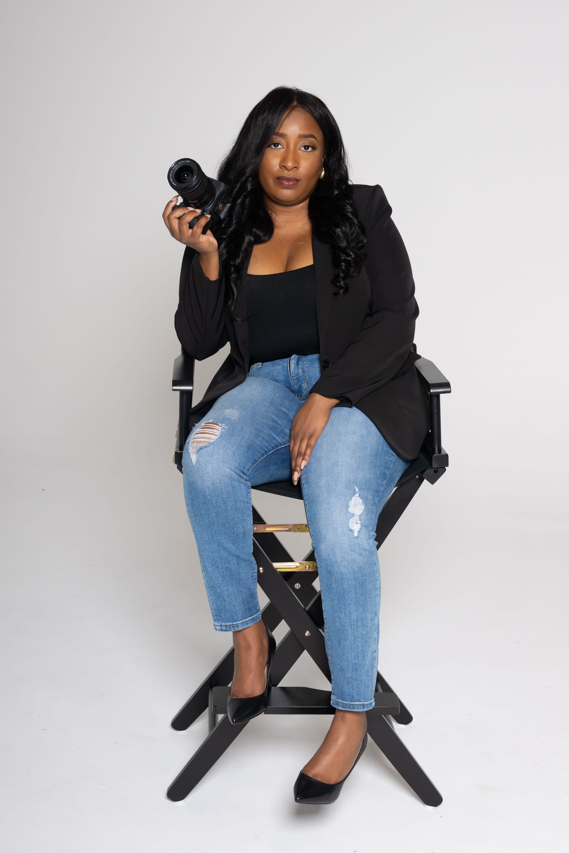 Woman seated on a director’s chair, holding a camera, wearing a black jacket, jeans, and heels against a plain backdrop