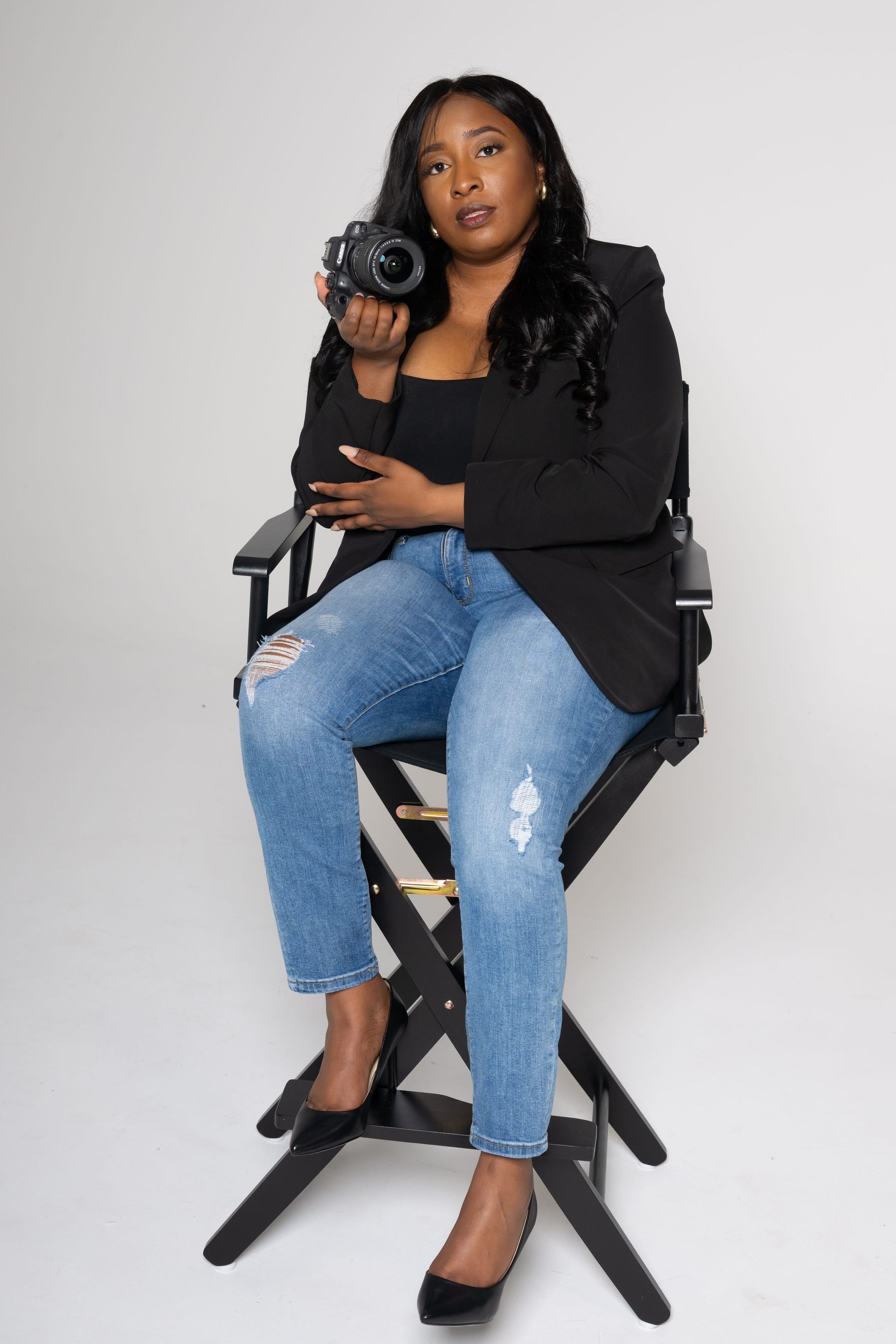 Woman seated on a director’s chair holding a camera, wearing black top and blue jeans against a light background