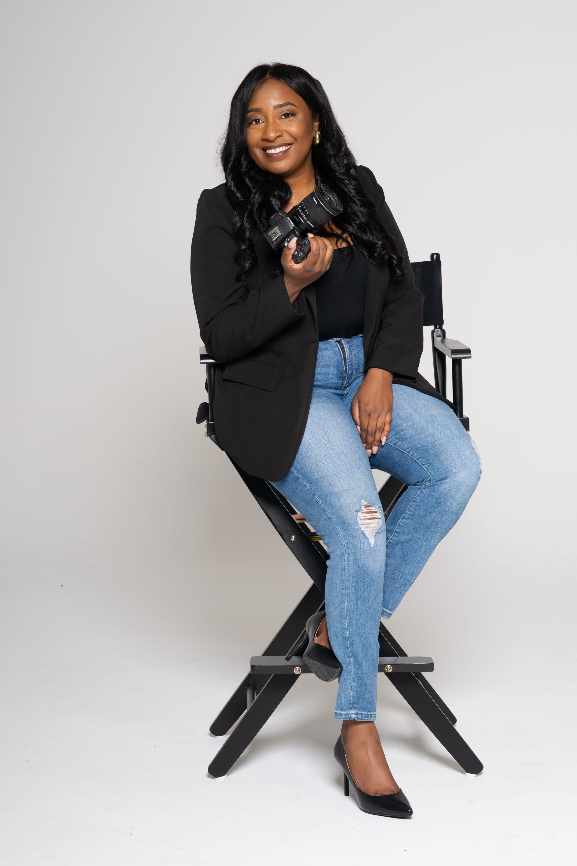 Woman seated on a director’s chair, smiling in a black blazer, jeans, and heels against a light gray backdrop