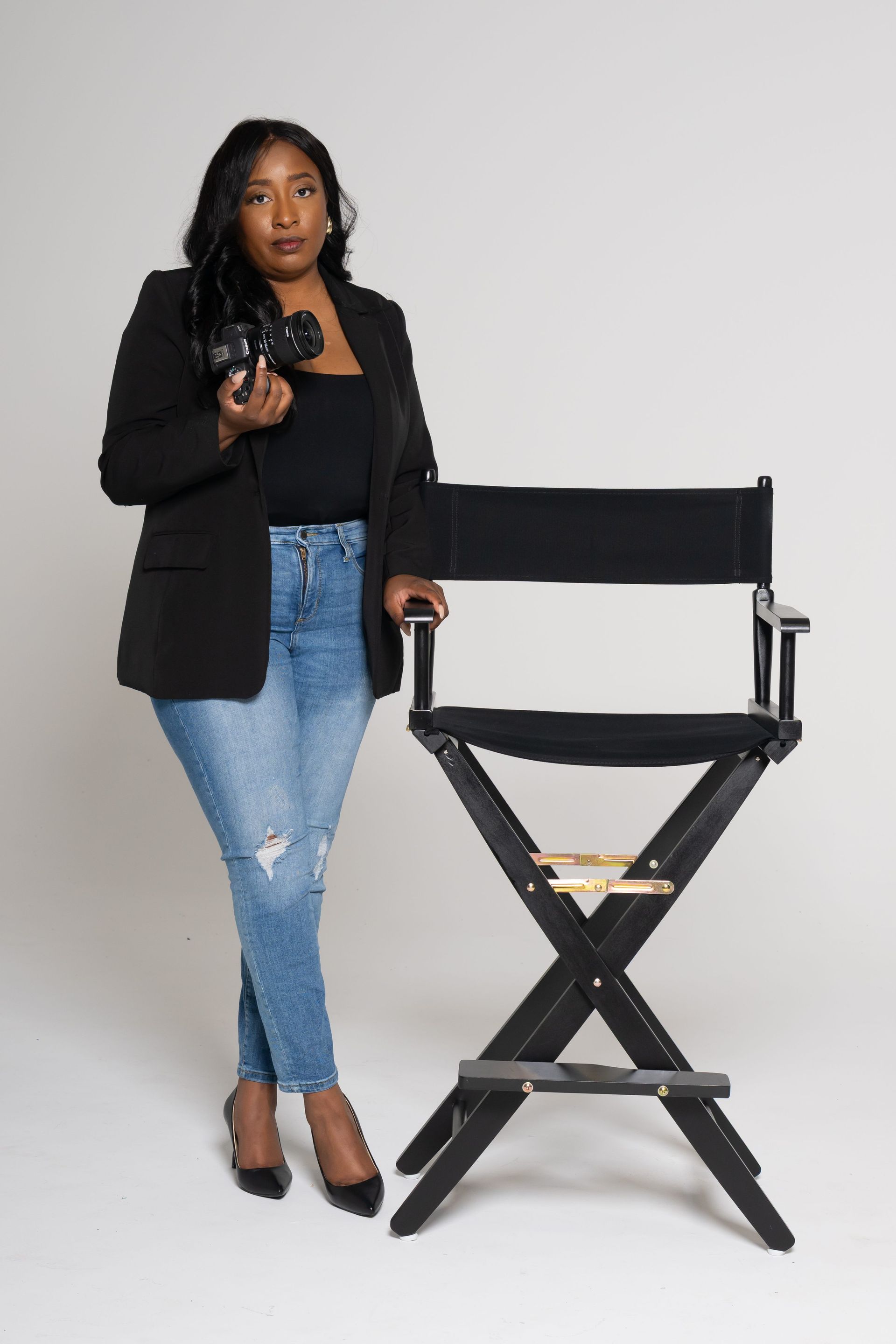 Woman in black blazer and jeans standing beside a black director’s chair, holding a camera in a studio.