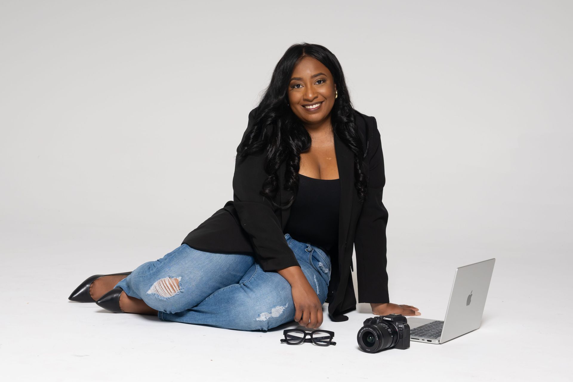 Woman in black jacket and jeans posing on a white backdrop beside a laptop, camera, and glasses