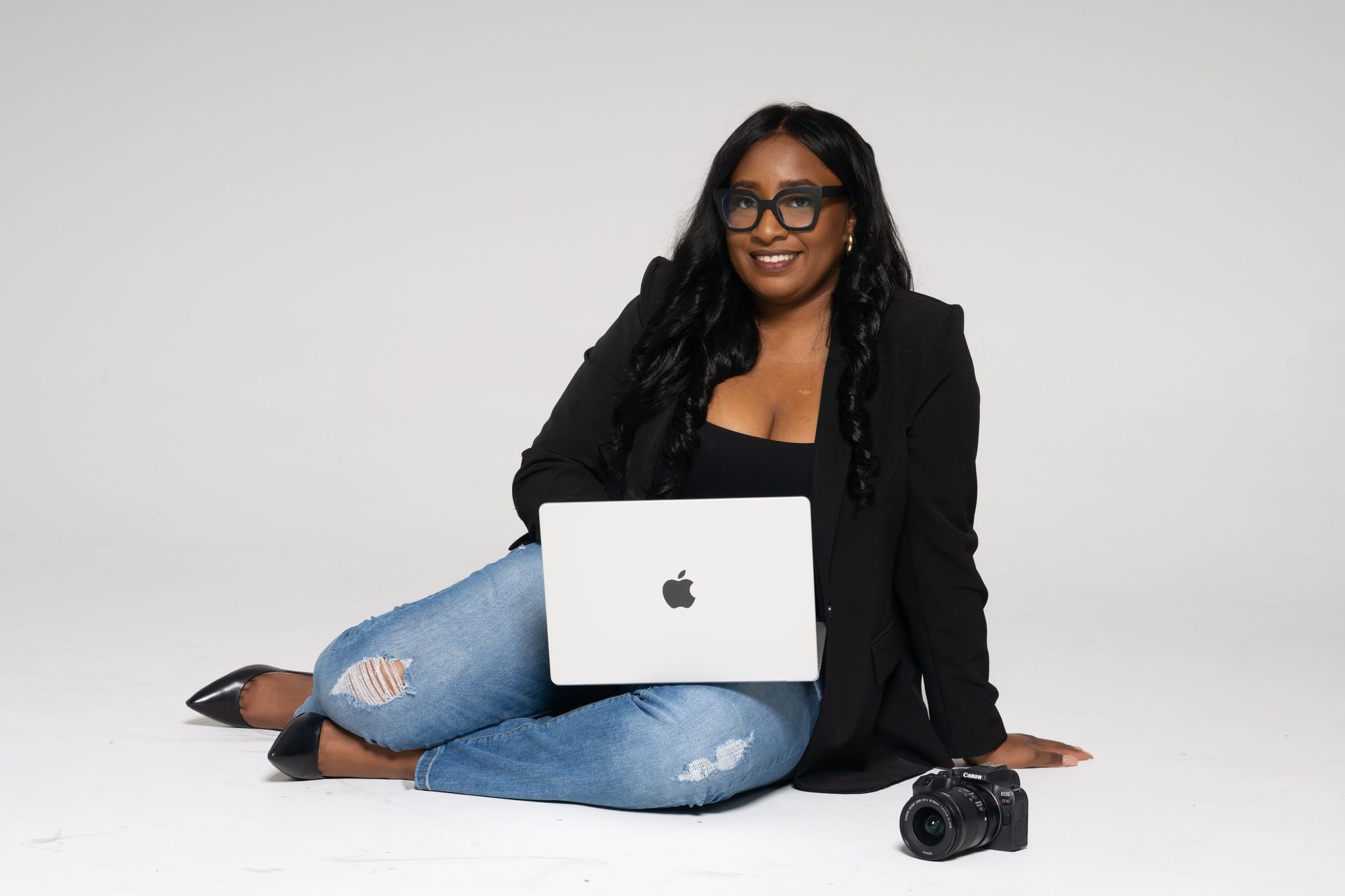 Woman seated with a silver laptop and camera on a white background, smiling at the camera.