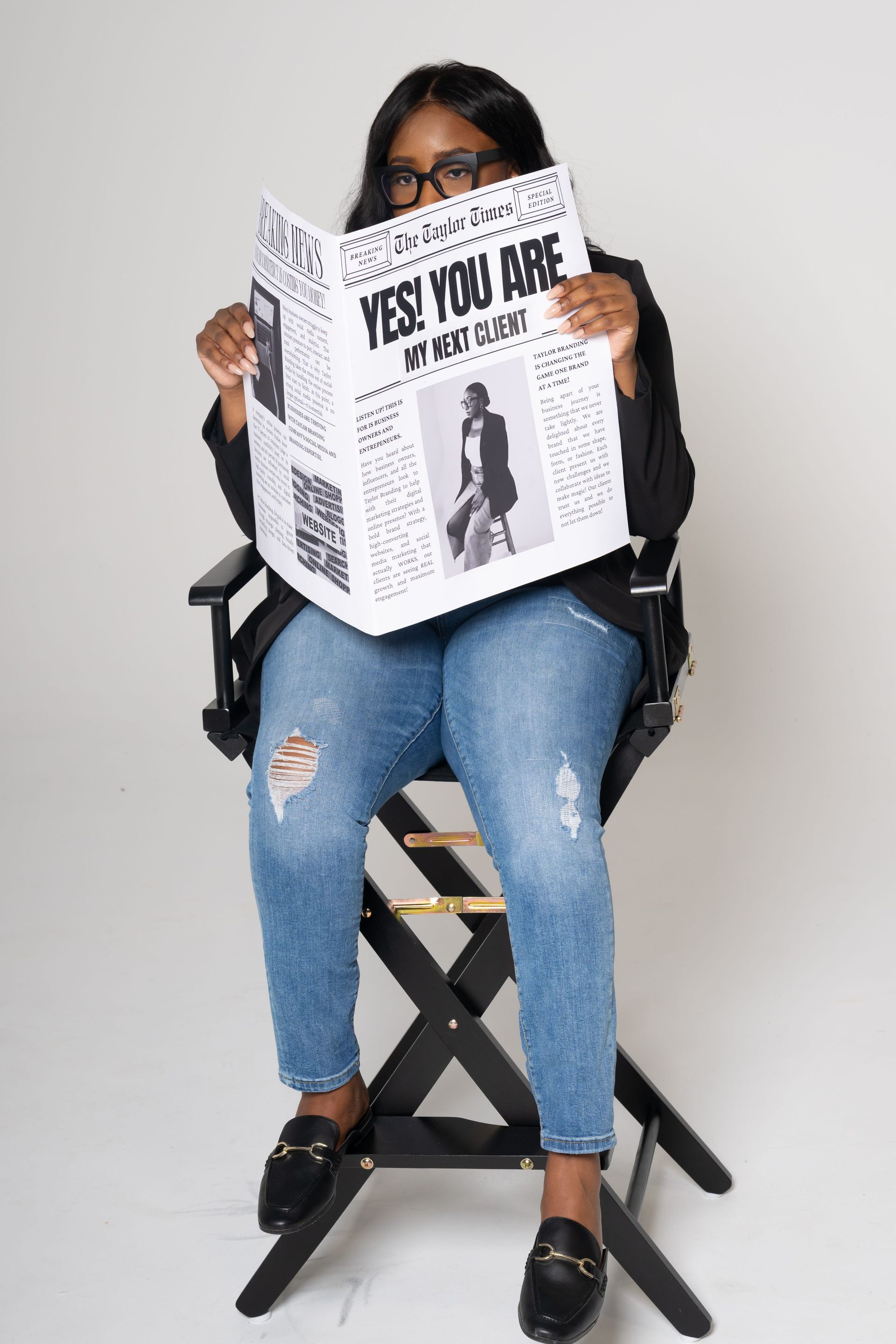 Person seated in a director’s chair reading a newspaper, wearing jeans and black shoes on a plain backdrop.