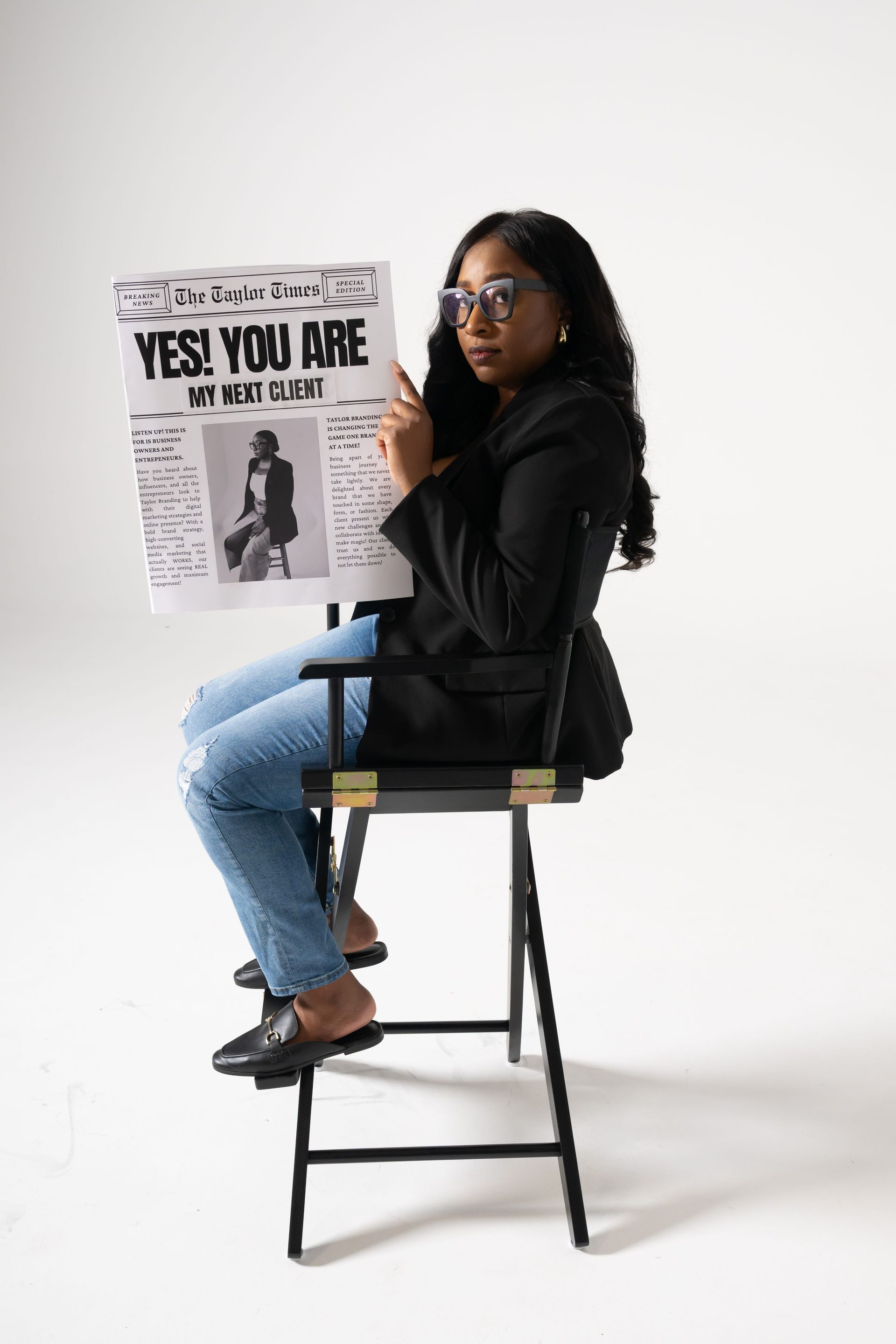 Woman seated on a tall stool holding a magazine, wearing black top, jeans, and boots against a white backdrop