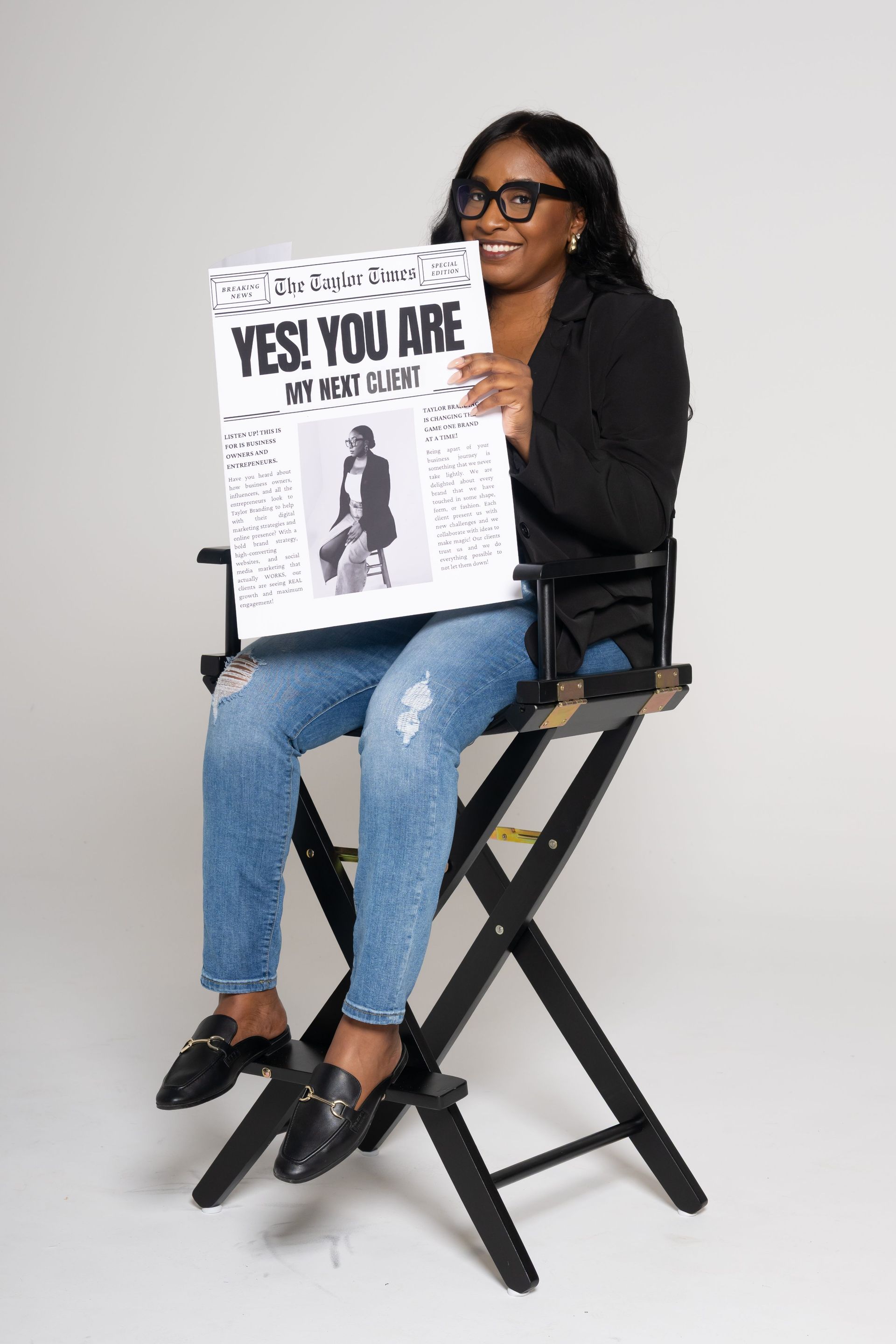 Person sitting on a director’s chair holding a “YES! YOU ARE” sign against a plain gray background
