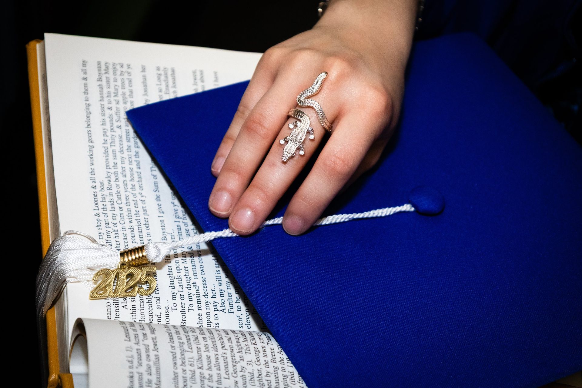 Hand with ornate ring resting on a blue diploma folder beside a rolled document and open book