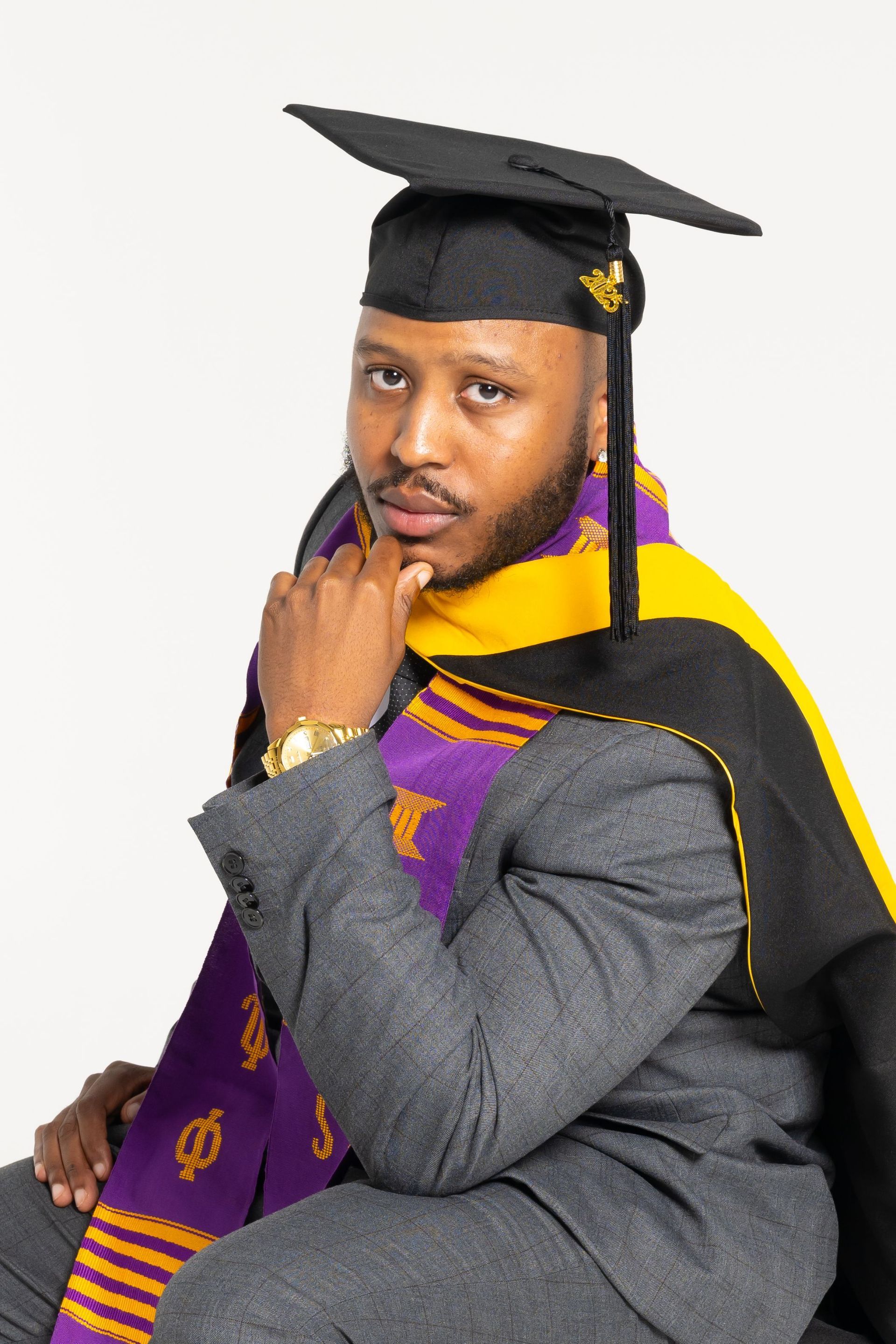 Graduation portrait of a person in cap and gown, posing with hand on chin against a plain background