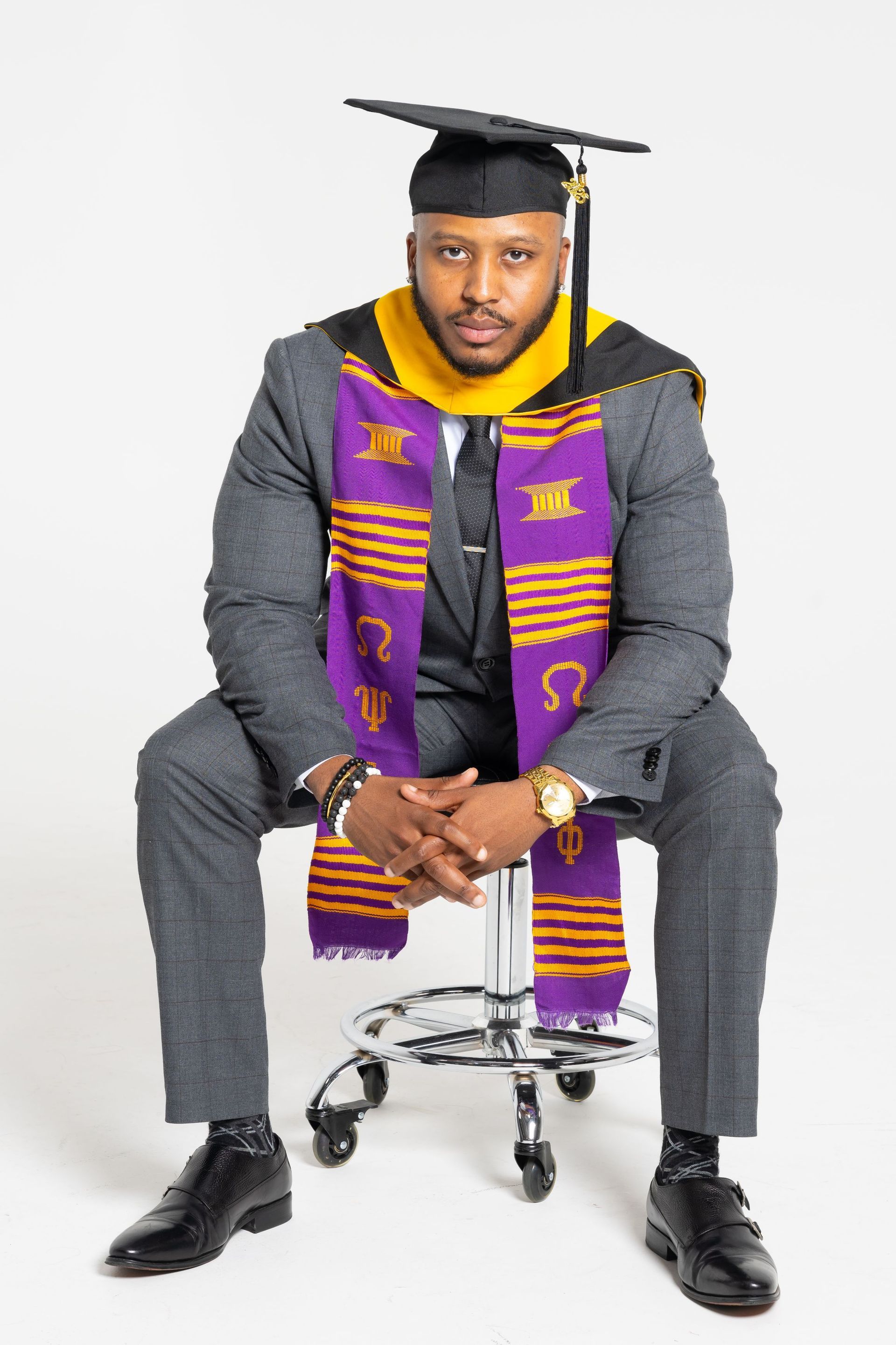 Graduation portrait of a seated graduate in gray suit with purple stole and cap on white background