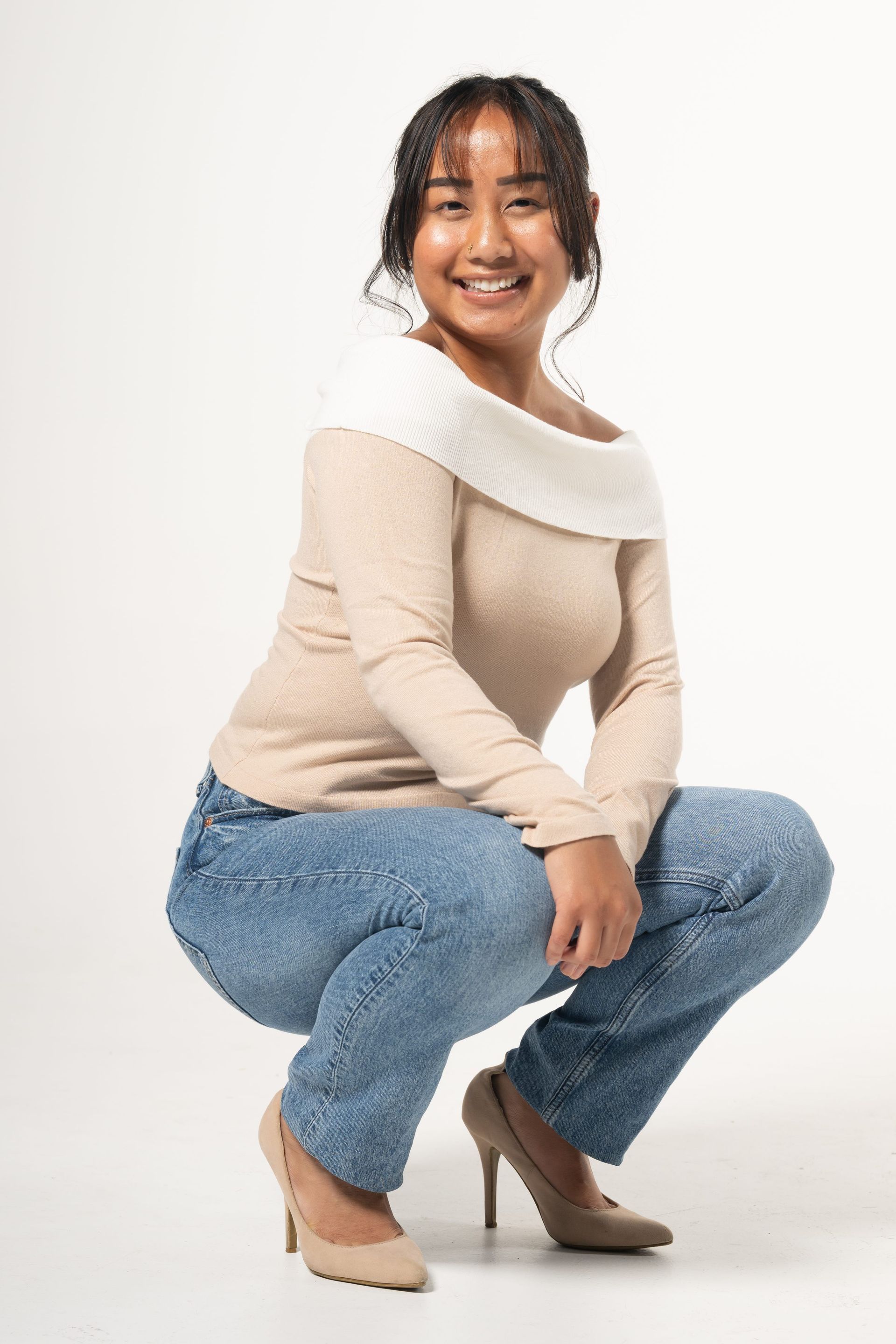 Smiling person crouching in a beige sweater, blue jeans, and heeled boots against a white background