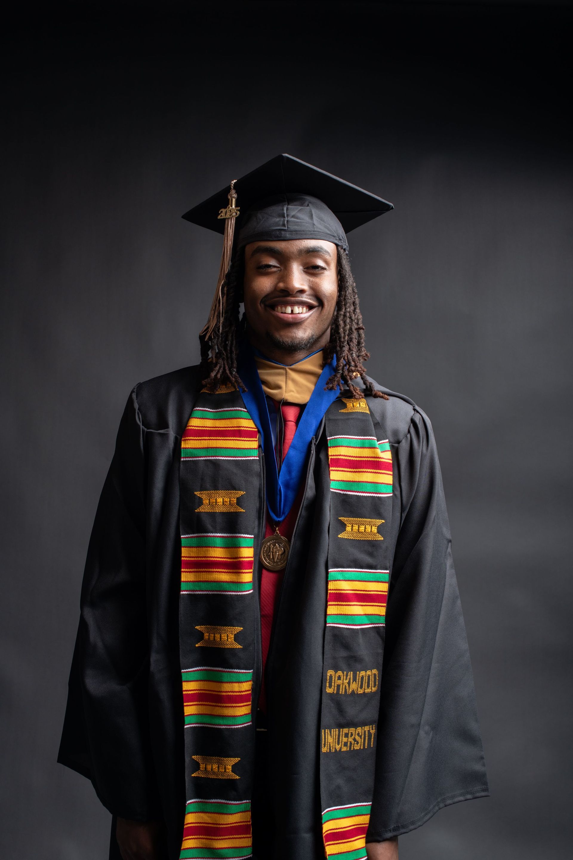 Graduate in cap and gown with colorful stole, smiling against a dark studio backdrop