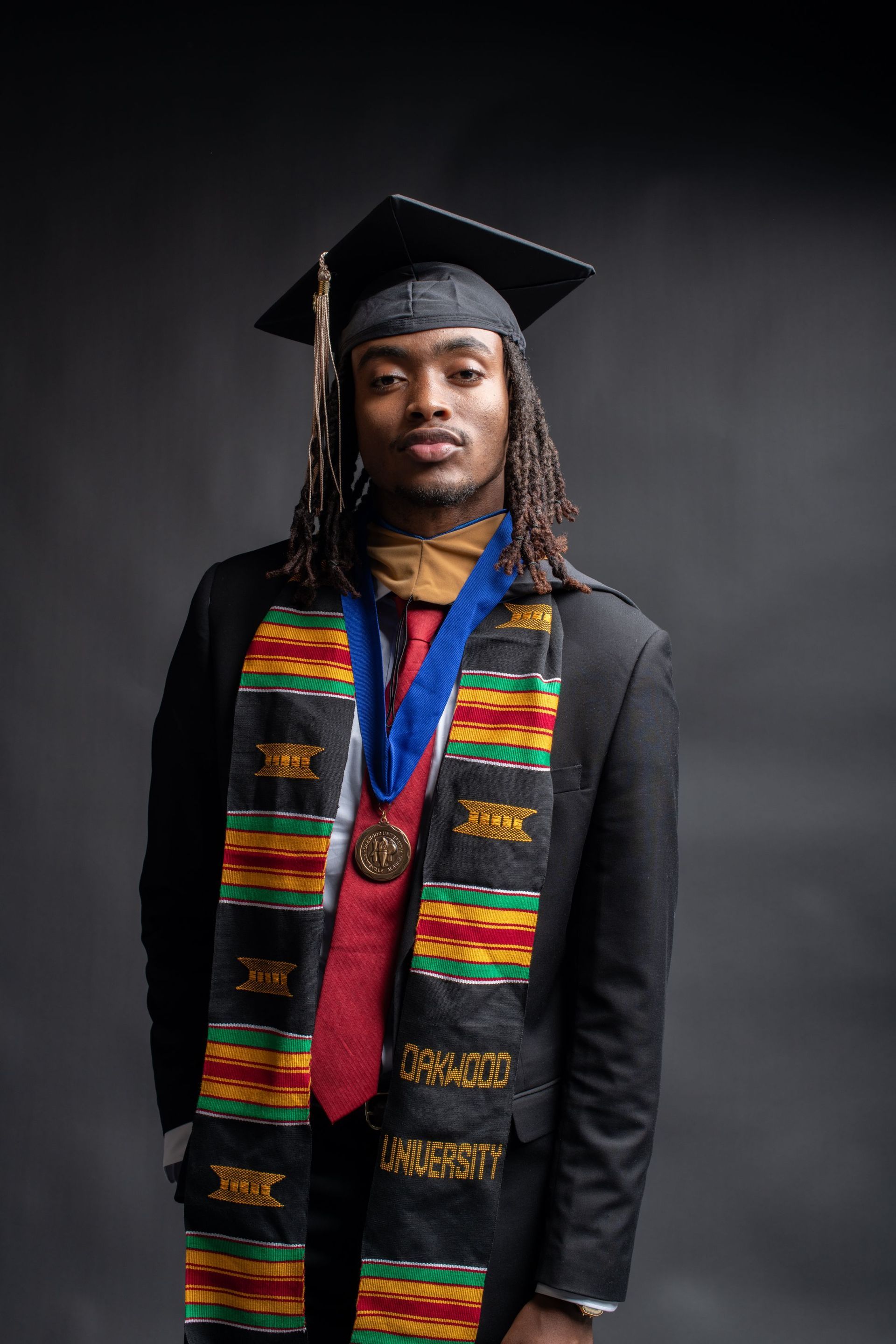 Graduate in cap and gown with colorful kente stole, standing against a dark studio background