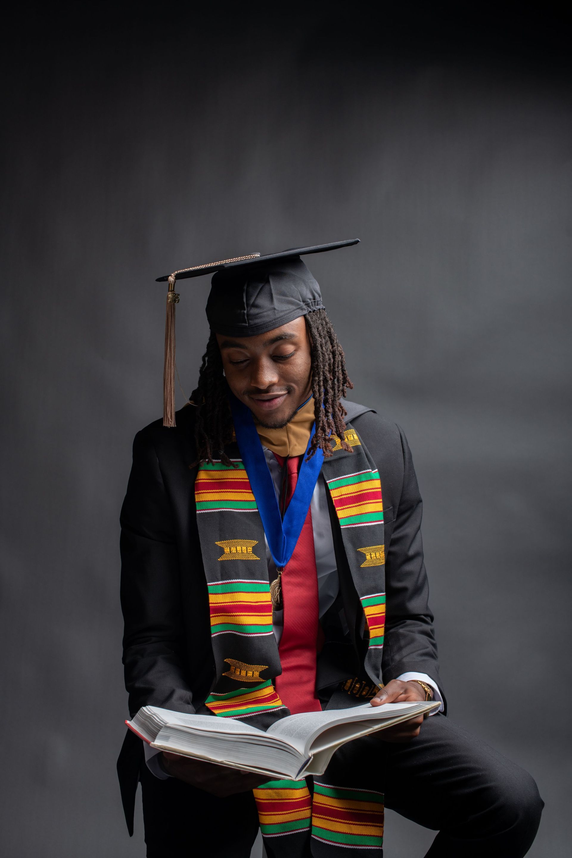 Graduate in black cap and gown with colorful stole, smiling while reading an open book against a dark backdrop