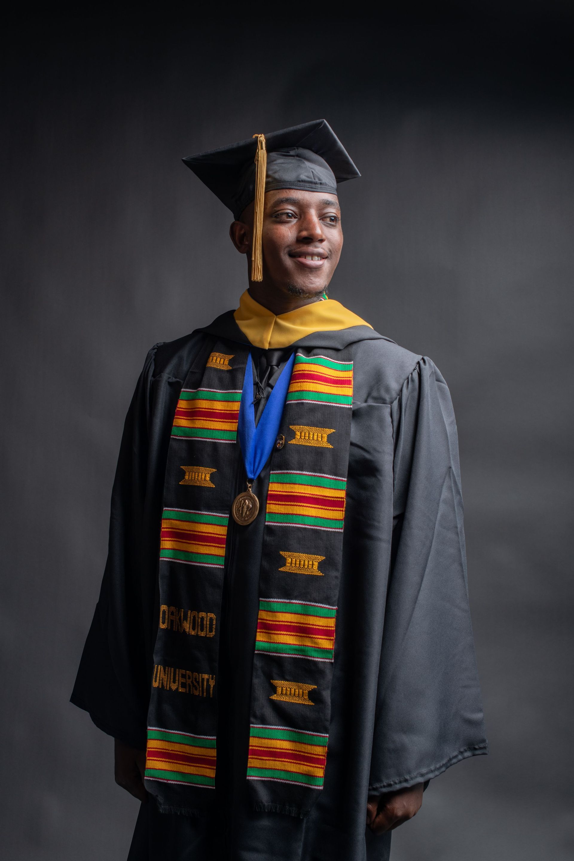 Person in graduation cap and gown with colorful stole, standing against a dark background.