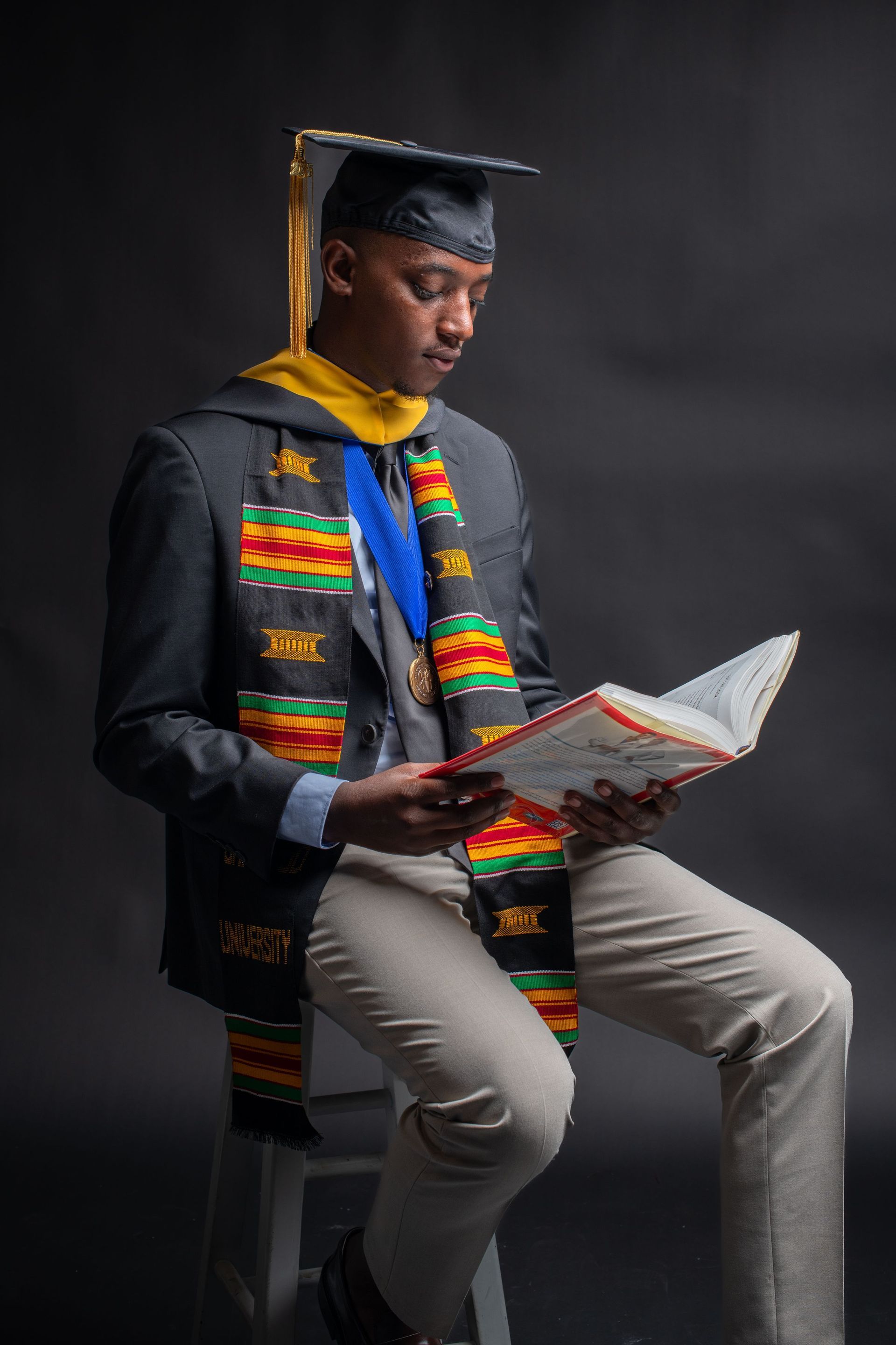 Graduate in cap and gown reading an open book against a dark studio backdrop