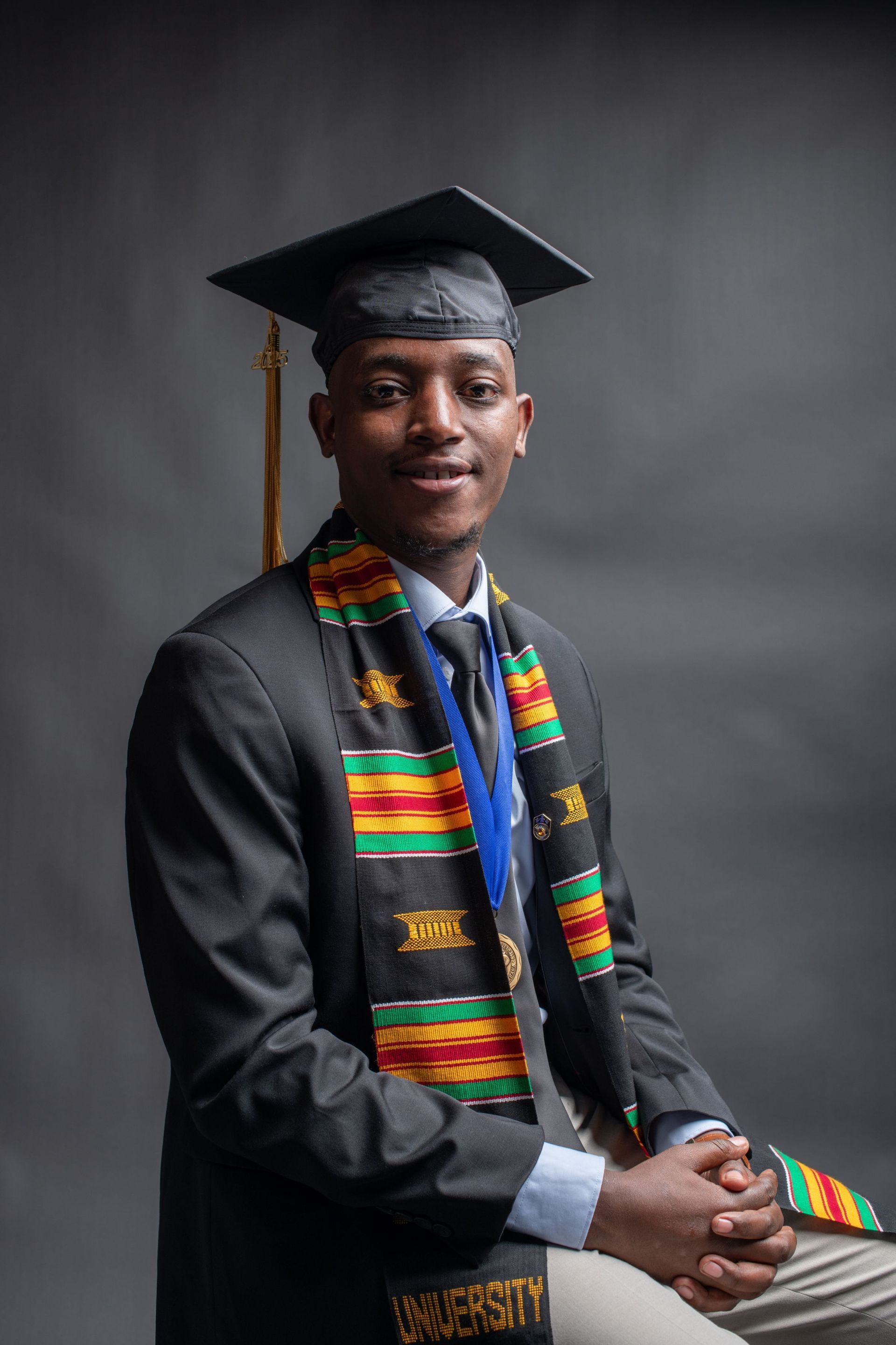 Graduate in cap and gown with colorful kente stole, seated against a dark studio background