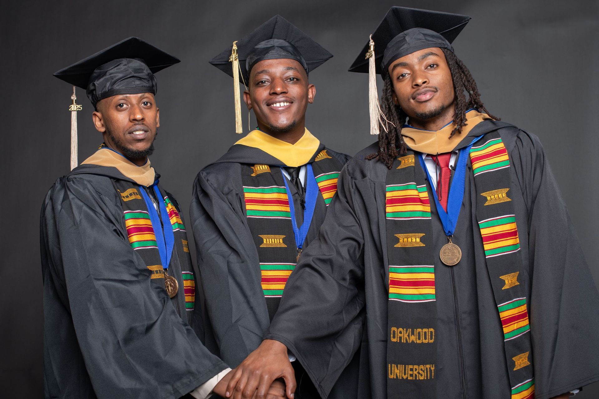 Three graduates in black caps and gowns pose together, smiling, with colorful stoles and medals.