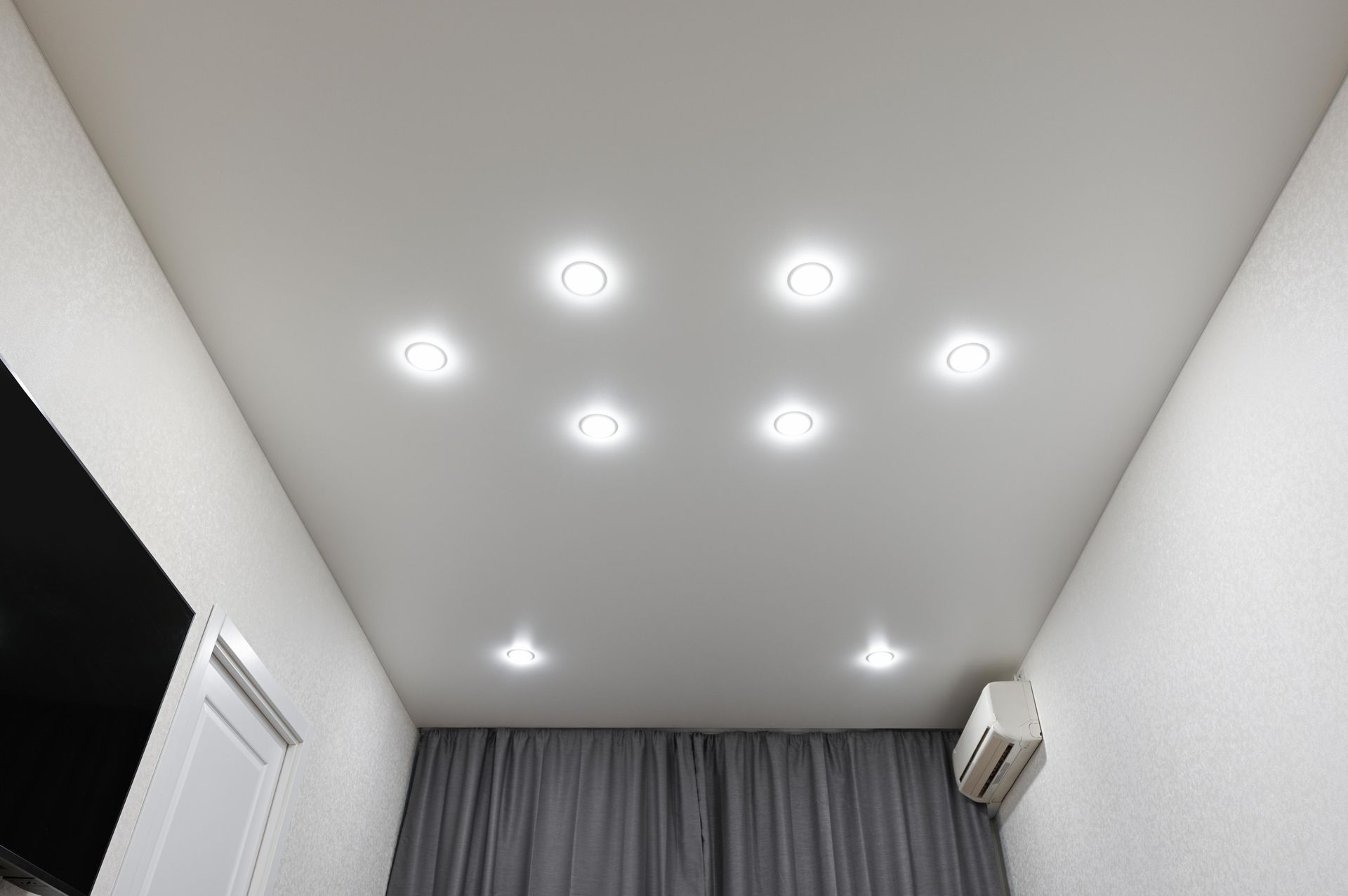 Ceiling with seven recessed lights, white walls, and a curtained window.
