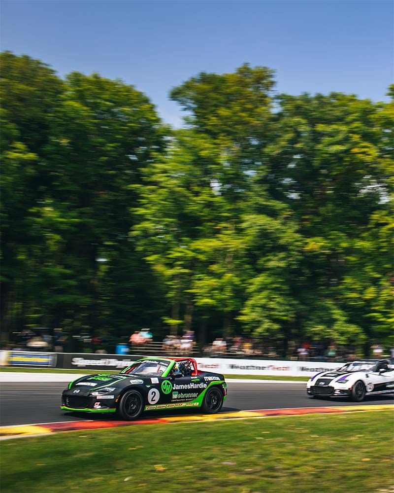 Two Mazda MX-5s are racing on a track with trees in the background.