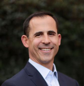 Smiling man in a dark suit and light blue shirt, outdoors with a green background.