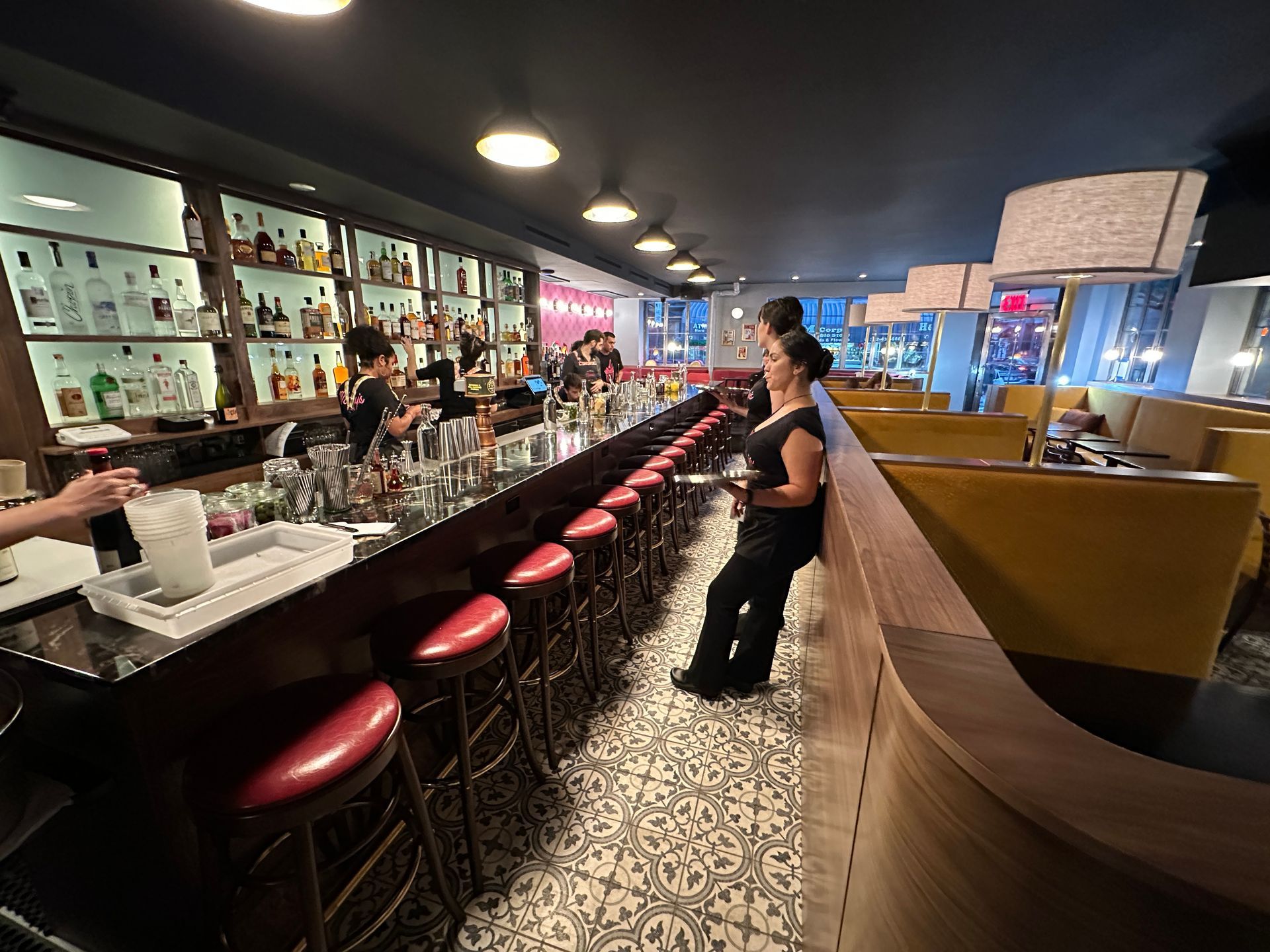 Modern cocktail bar with red stools, patterned floor, and a bartender behind the counter