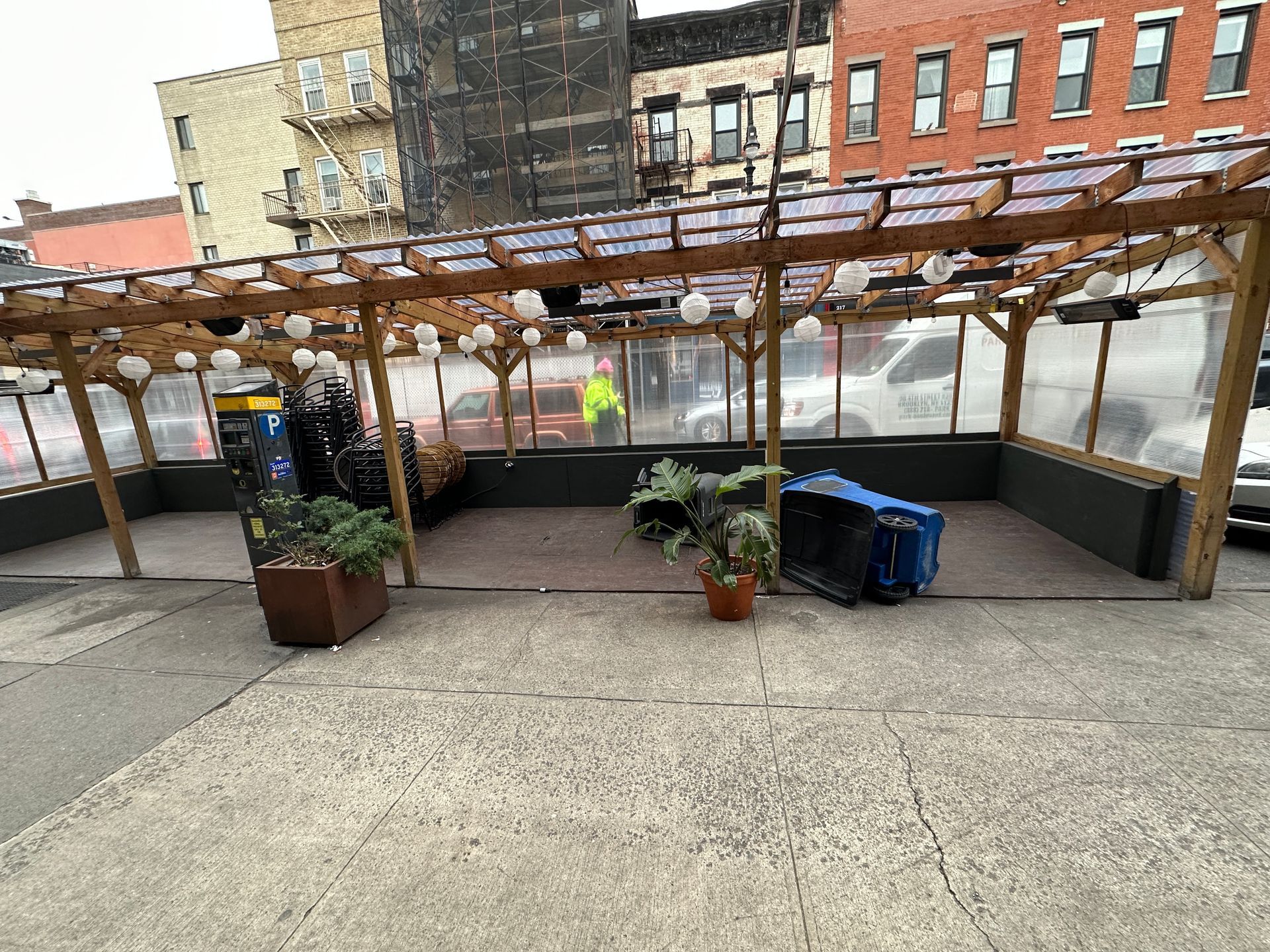 Outdoor patio with pergola, potted plants, and blue recycling bins beside a brick building.