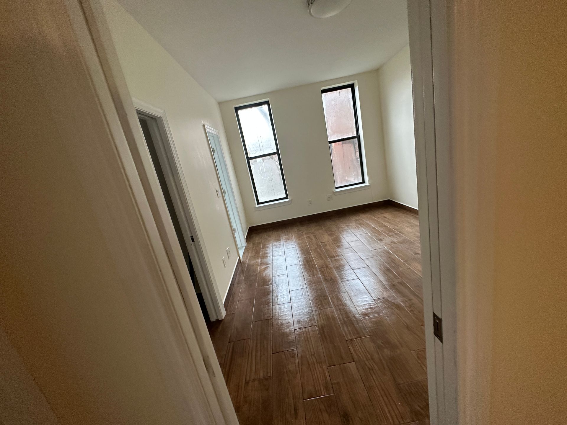 Empty unfurnished room with hardwood floors and two tall windows, viewed from the hallway