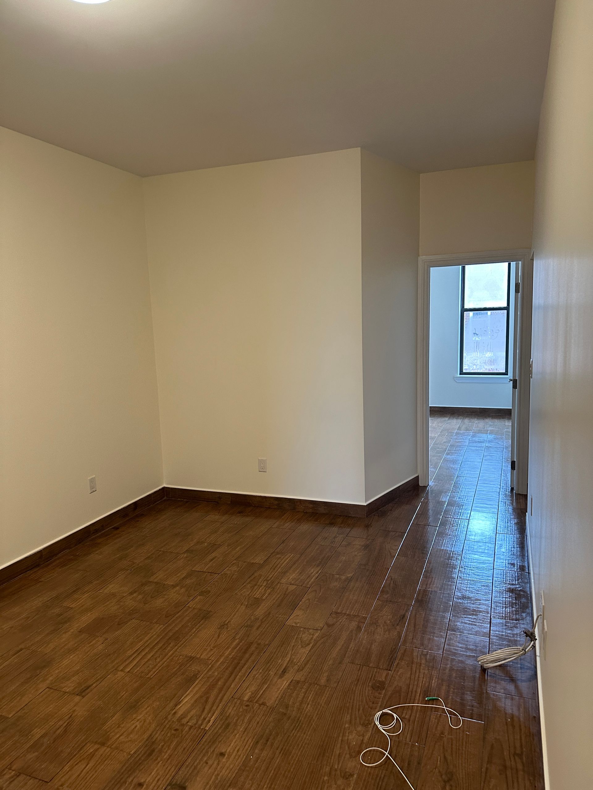Empty room with hardwood floors, beige walls, and a narrow hallway leading to a window.