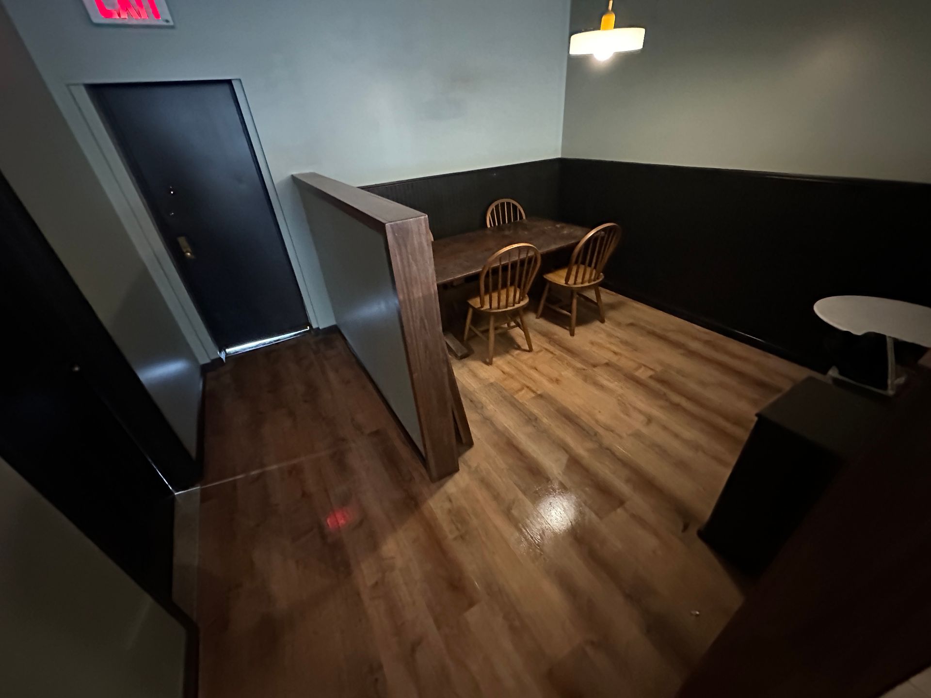 Small dining area with wooden floor, partition wall, two chairs, round table, and dark door by an exit sign