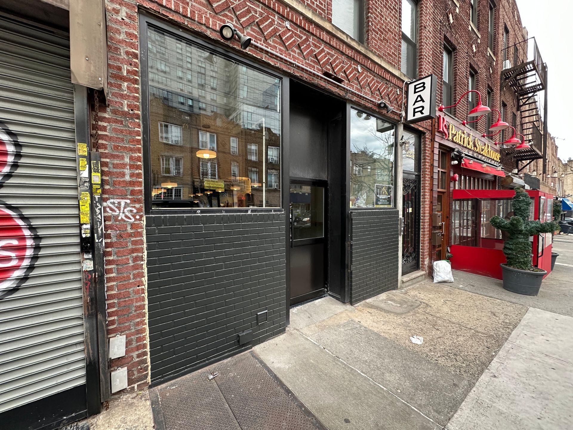 Brick storefront with black metal door and window on a city sidewalk, beside a red kiosk and shuttered storefront.