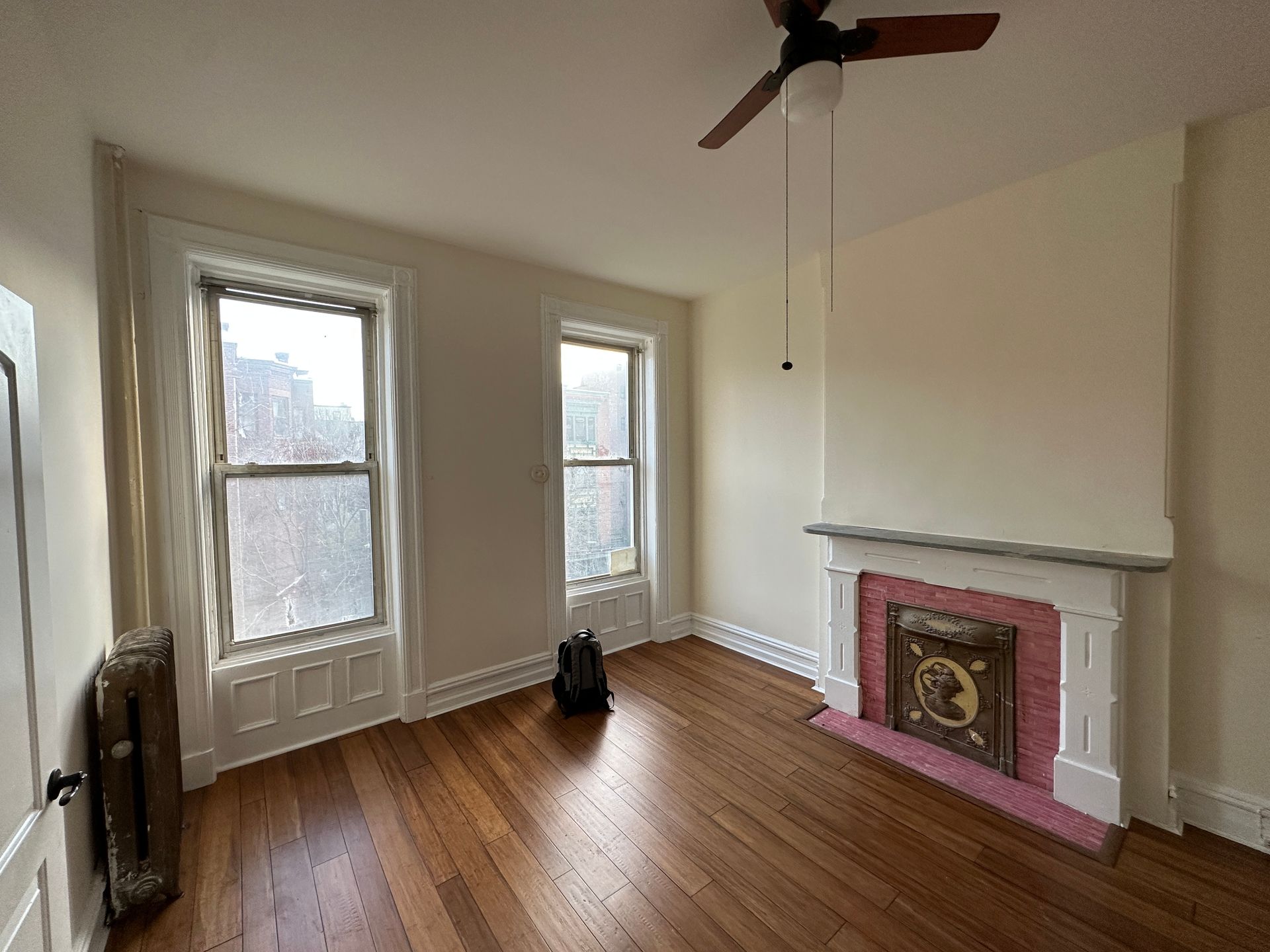 Empty room with hardwood floors, two tall windows, and a pink fireplace mantel under a ceiling fan.