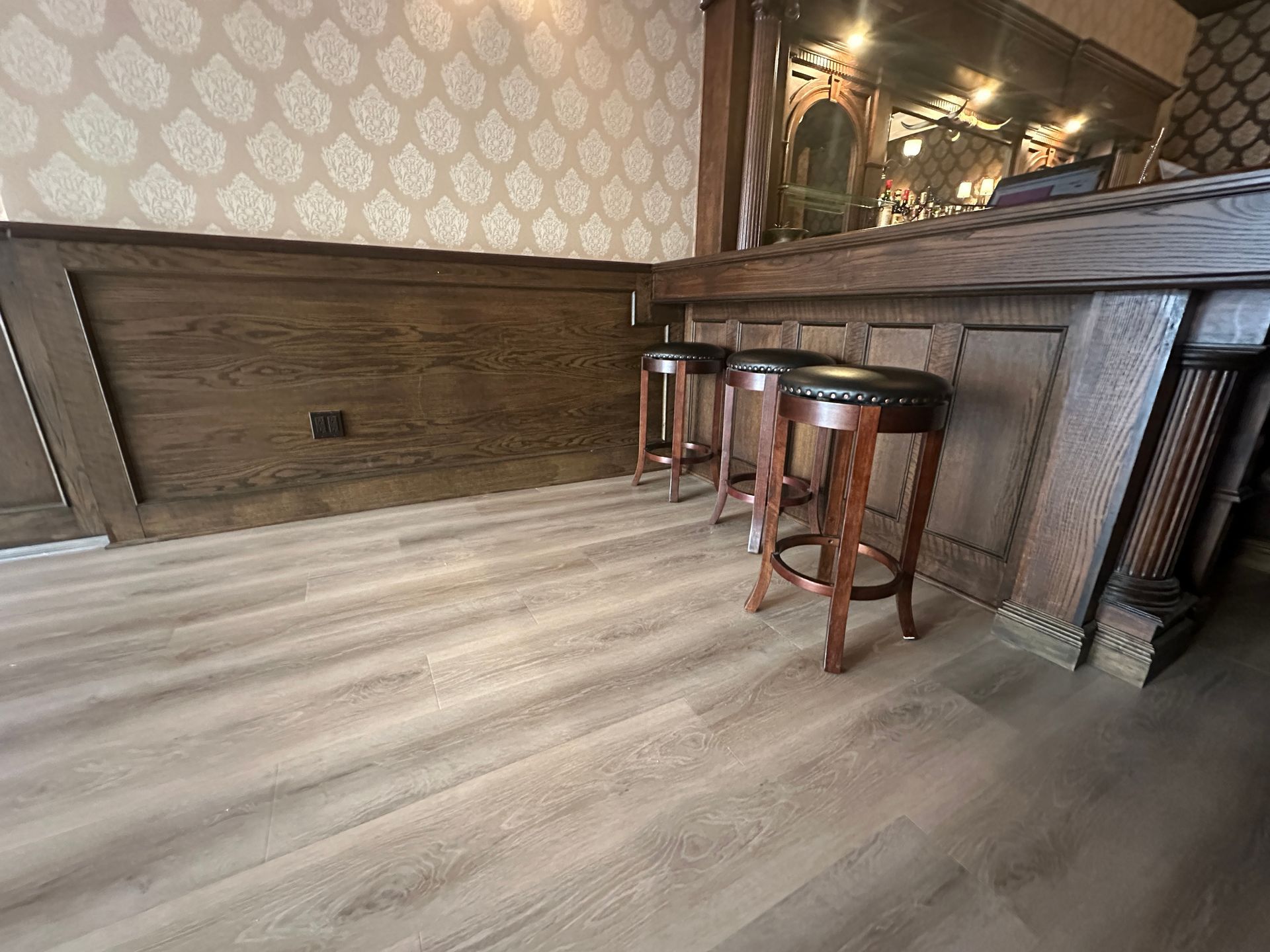 Empty bar area with three wooden stools, patterned wallpaper, and a dark wood counter on light flooring