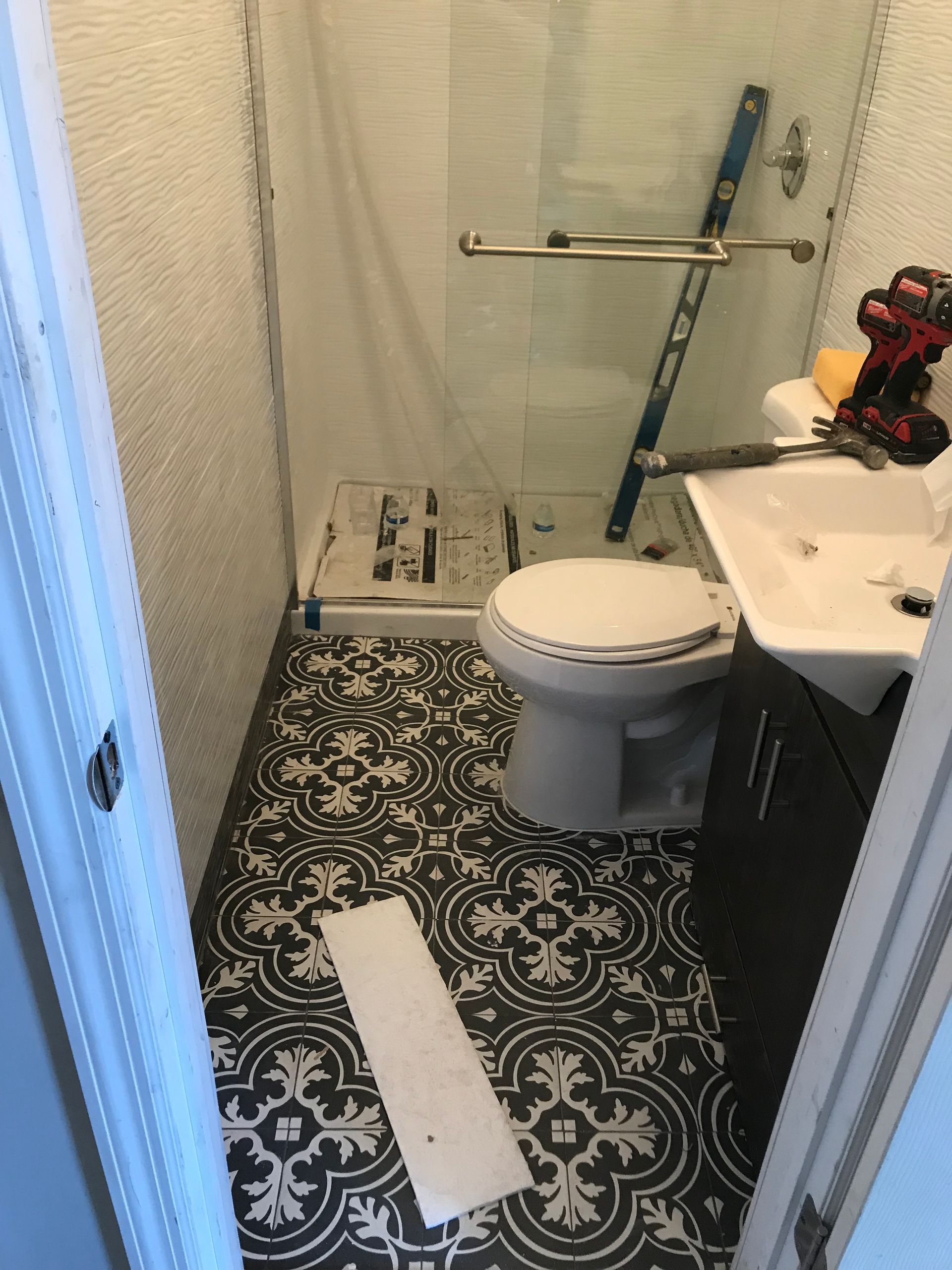 Small bathroom with toilet, sink, shower curtain, grab bars, and patterned floor mat.