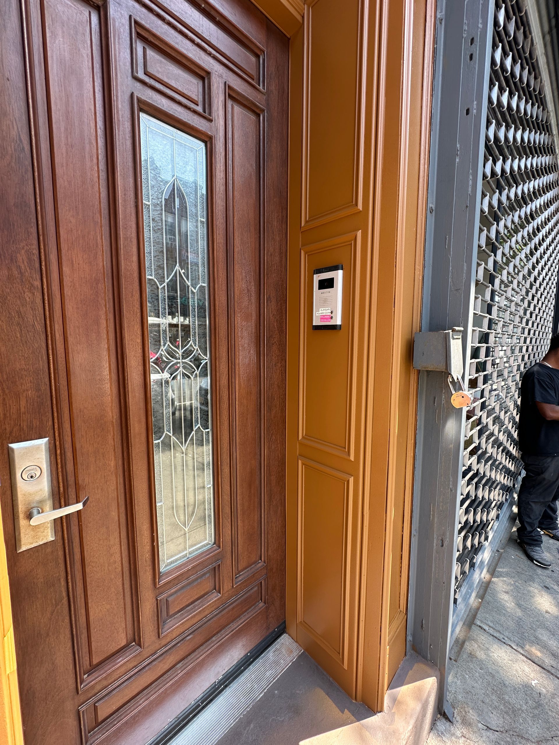 Wooden front door with glass panel beside a metal security gate and intercom on a brick entryway