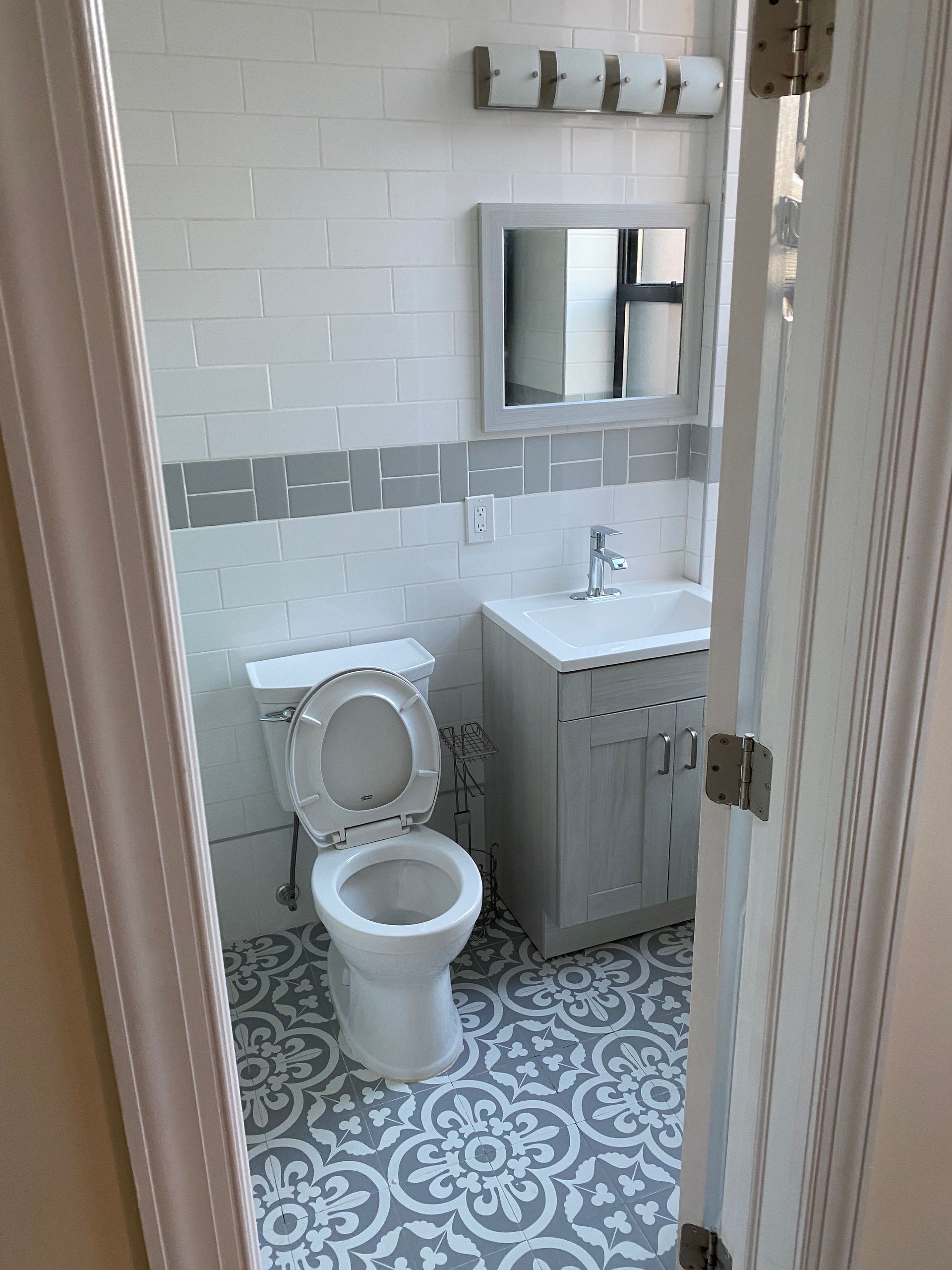 Small white bathroom with toilet, sink cabinet, mirror, and patterned blue floor tile.
