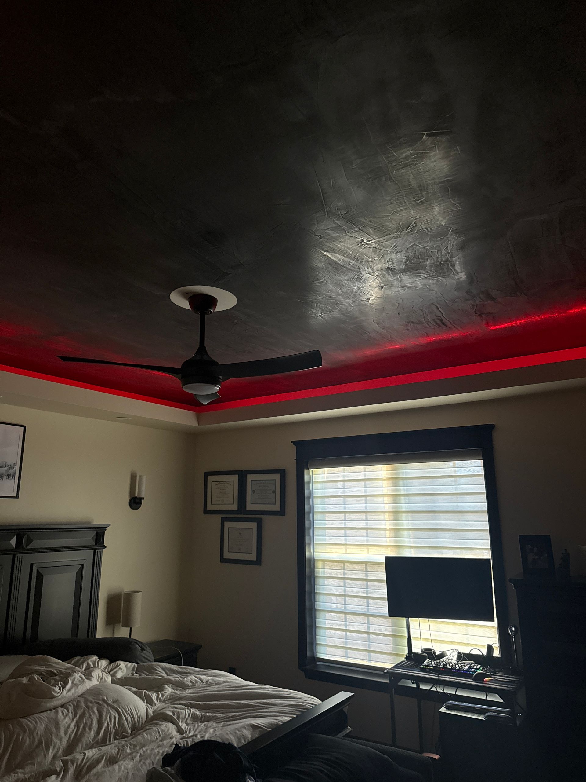 Dim bedroom with unmade bed, ceiling fan, red LED light strip, window blinds, and wall decor.